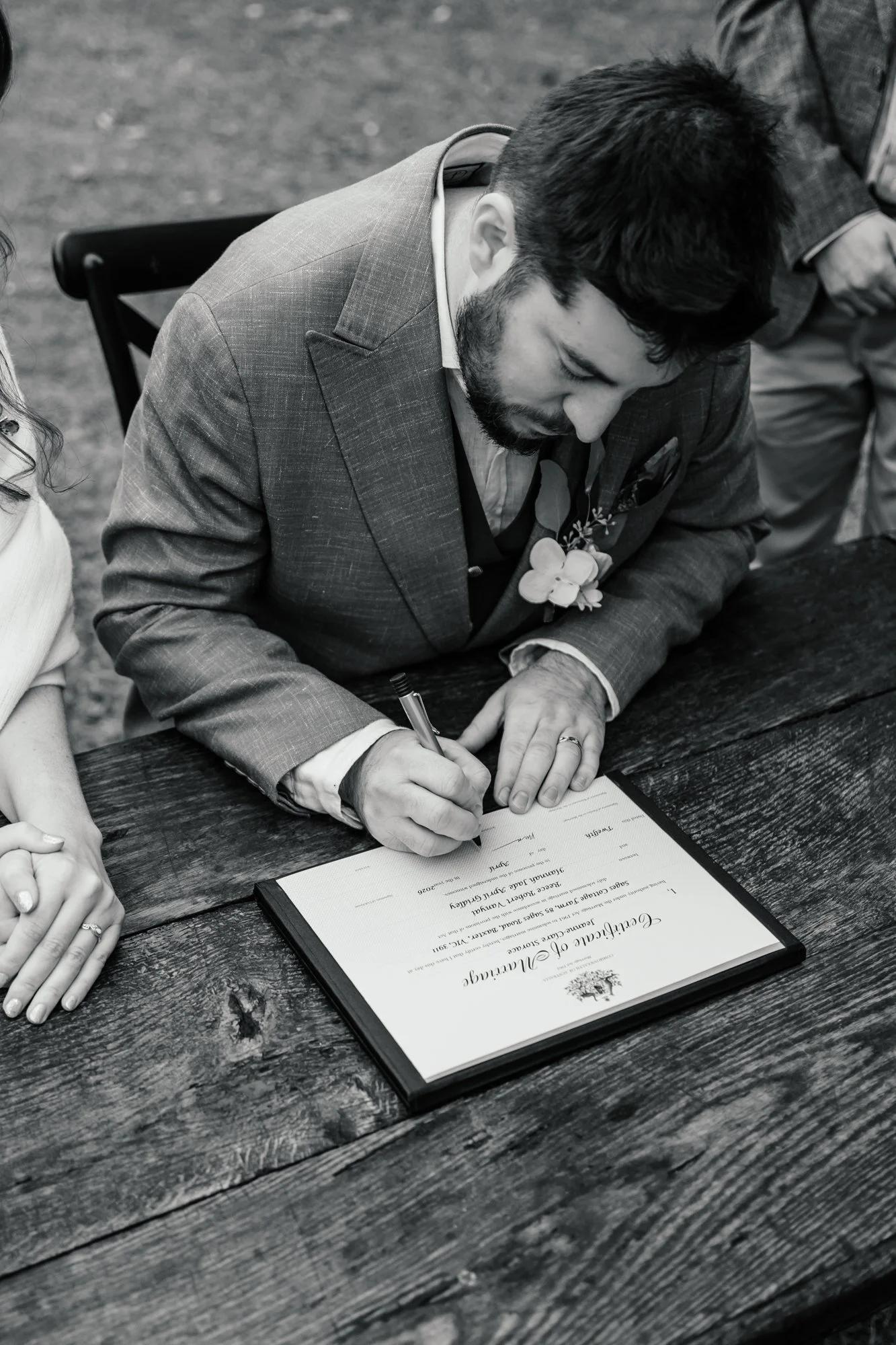 A man in a suit signing a certificate on a wooden table during a formal outdoor event.