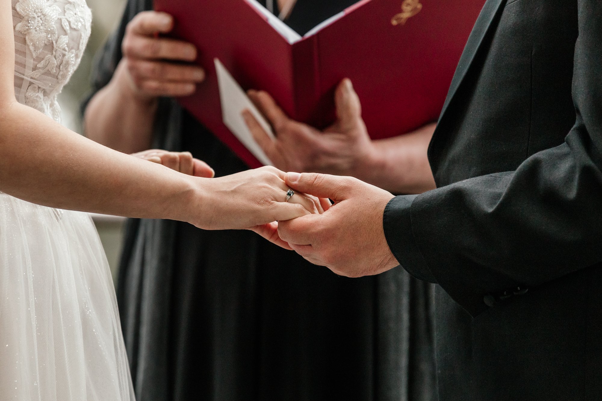 Close-up of a wedding ceremony with a bride and groom holding hands, the bride's hand has a ring, and an officiant in the background holding a red book.