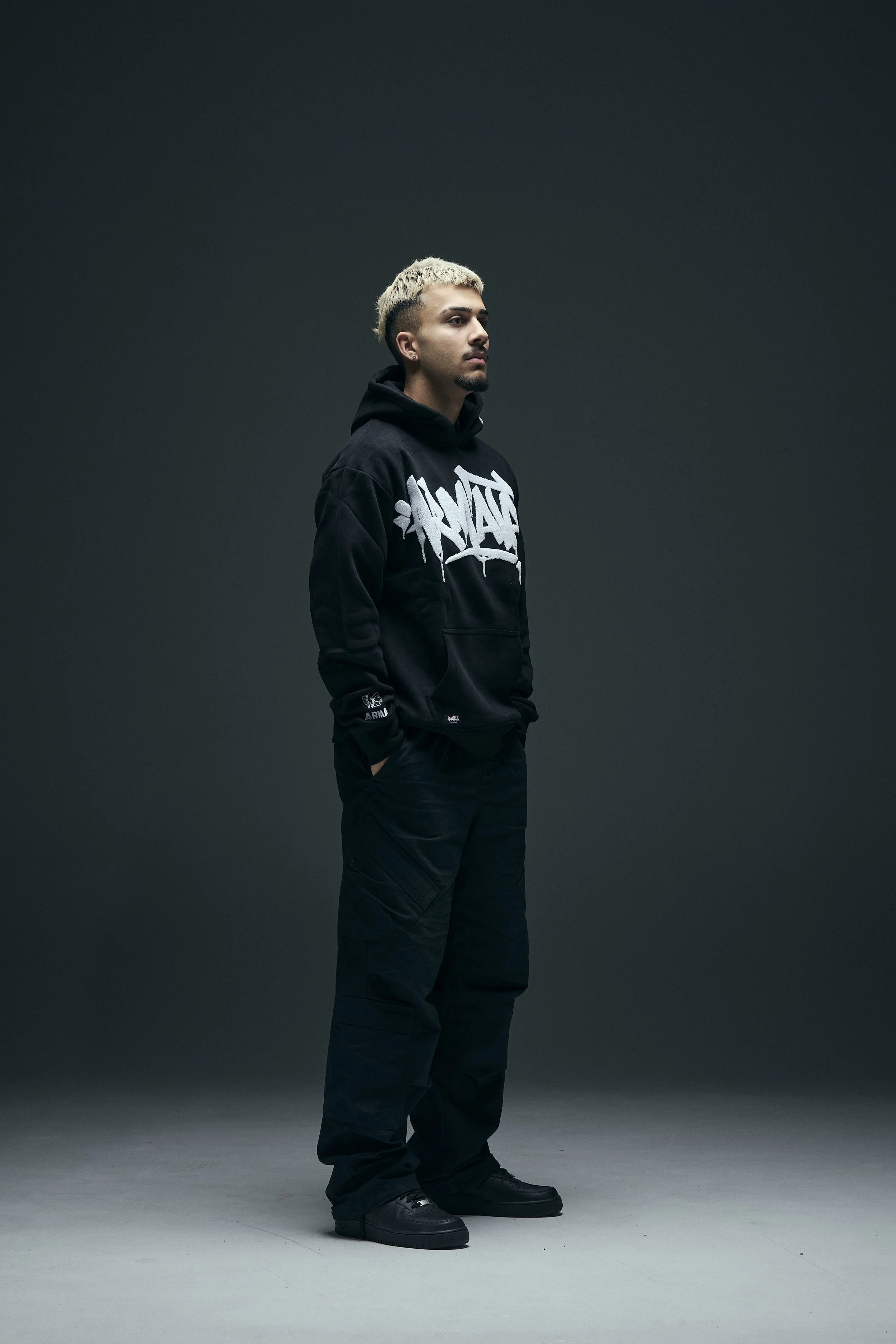 A young man with blond hair and a goatee is standing in profile against a dark gradient background. He is wearing a black hoodie with graffiti-style white text on the front and black pants, with his hands in his pockets.
