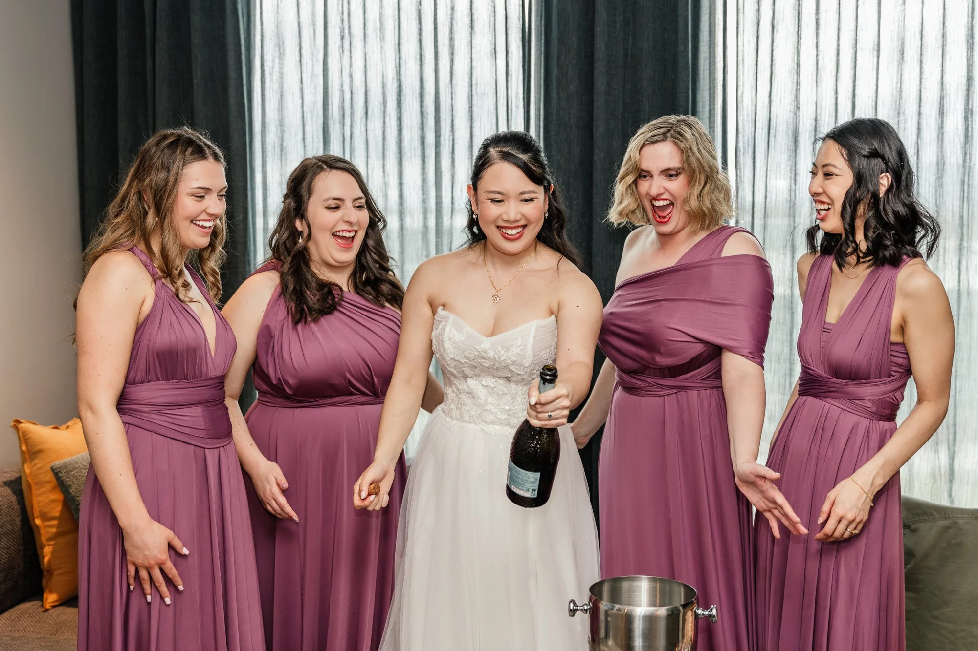 A bride in a white wedding dress holding a champagne bottle, surrounded by five bridesmaids in matching mauve dresses, all smiling and looking at the champagne.