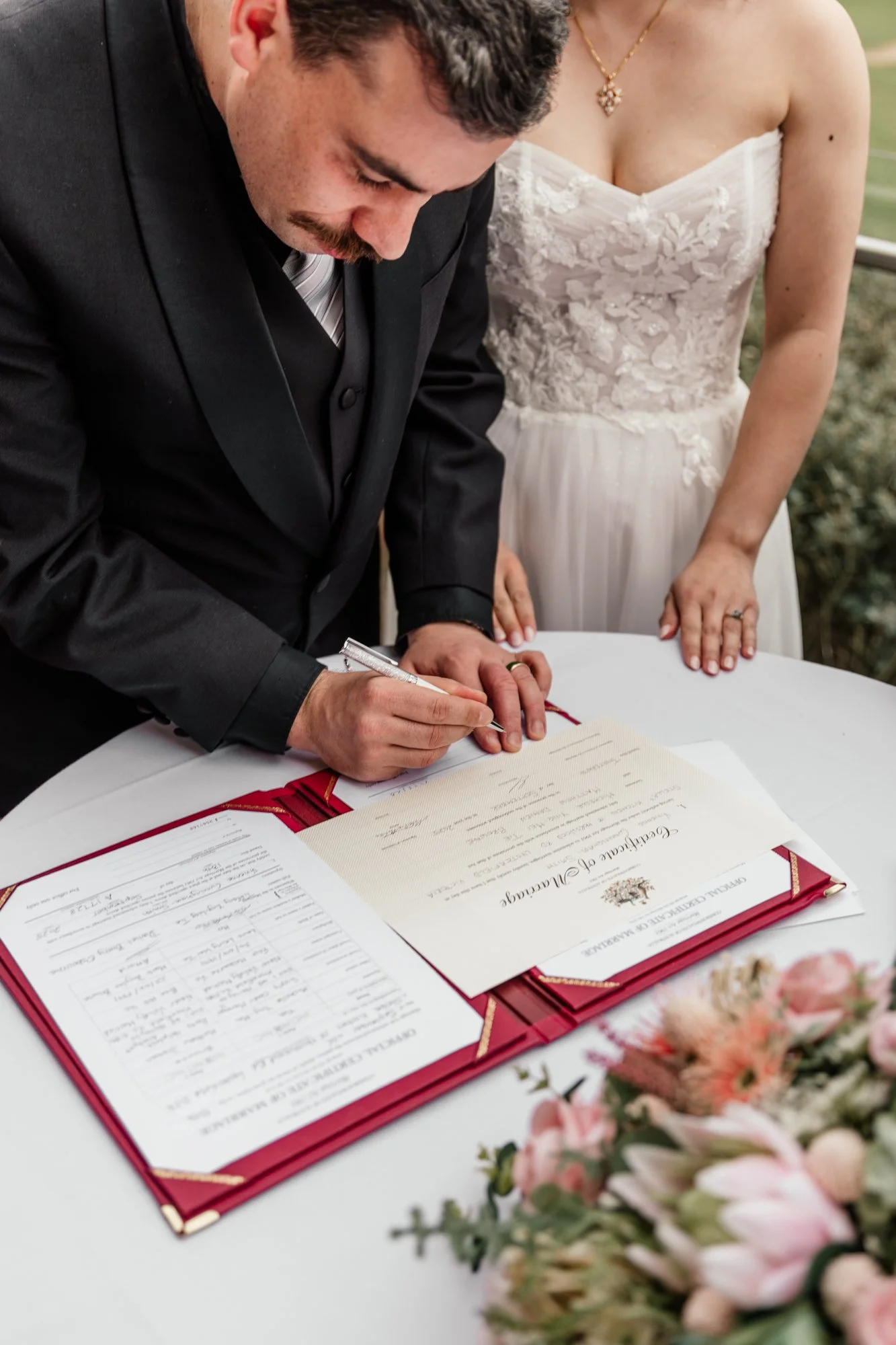 A man in a black suit signs a wedding marriage certificate with a gold wedding band on his finger, while a woman in a white lace wedding dress and a gold necklace stands beside him. There are documents and a bouquet of flowers on the table.