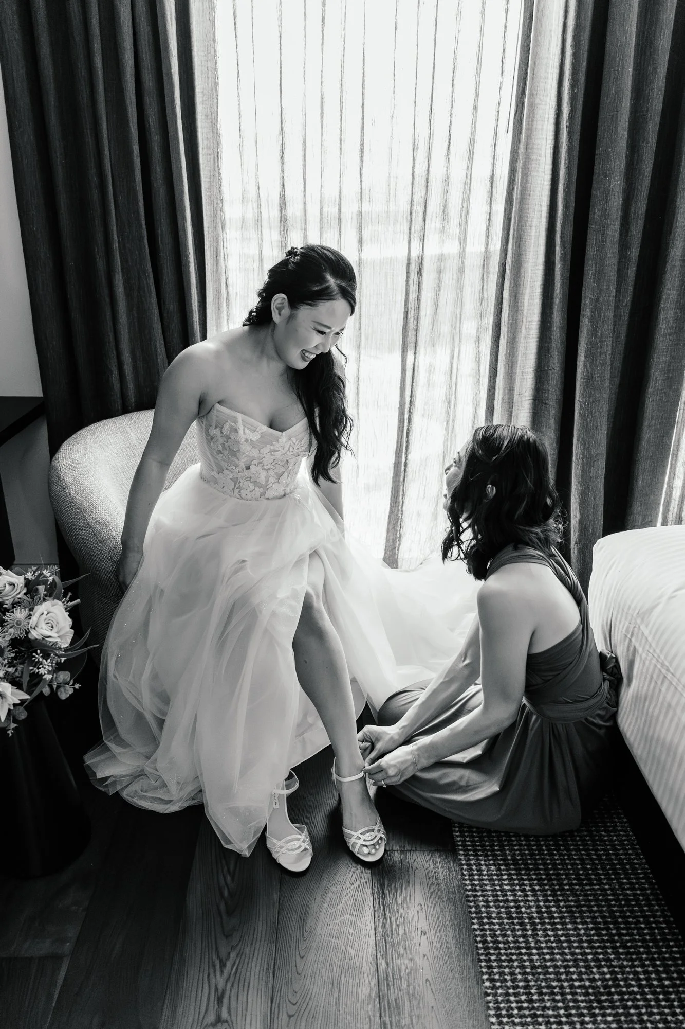 A bride in a strapless wedding dress smiling as a woman helps her put on her shoes in a hotel room. There are dark curtains and a window in the background, with flowers on a table to the side.