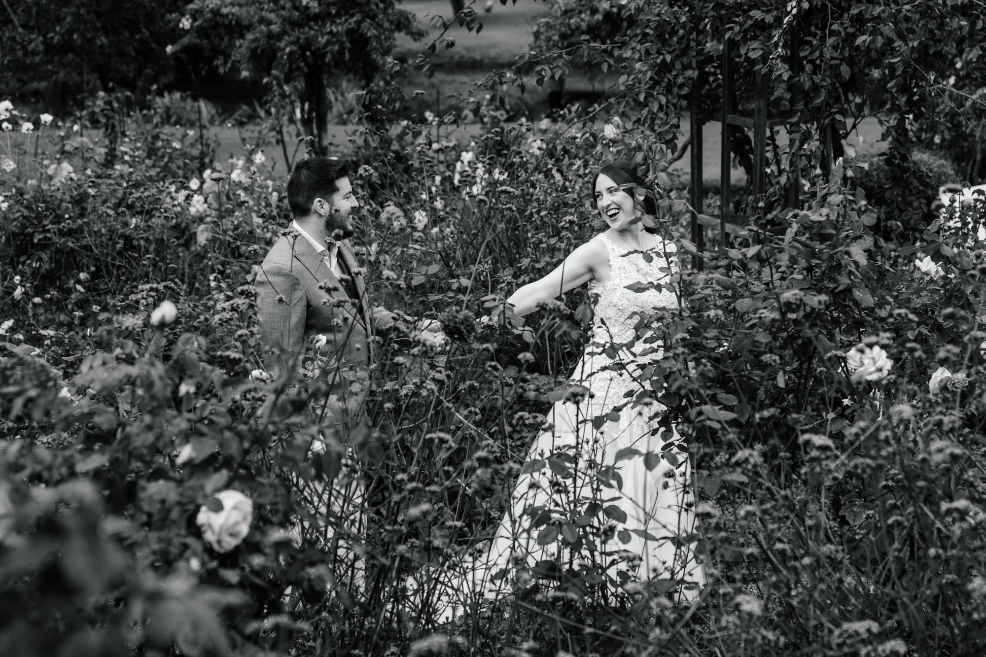 A black and white photograph of a man and a woman standing in a garden or park, surrounded by dense foliage and flowers. The man is wearing a suit, and the woman is dressed in a lace dress. They are smiling and looking at each other, appearing happy 