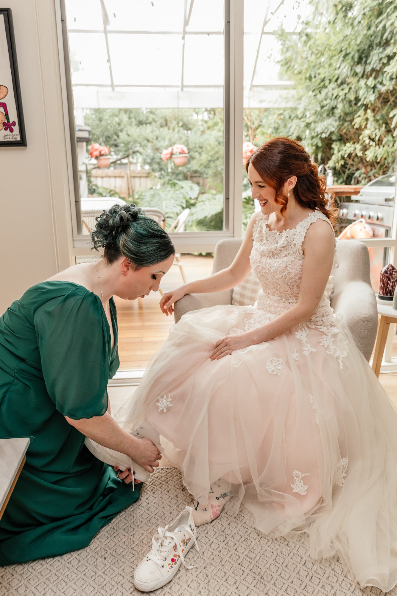 A bride is sitting on a beige chair, smiling, as her dress is being adjusted by a woman in a green dress. The woman is kneeling on the floor, putting on a white sneaker with floral details, while the bride wears a white lace wedding gown. They are in