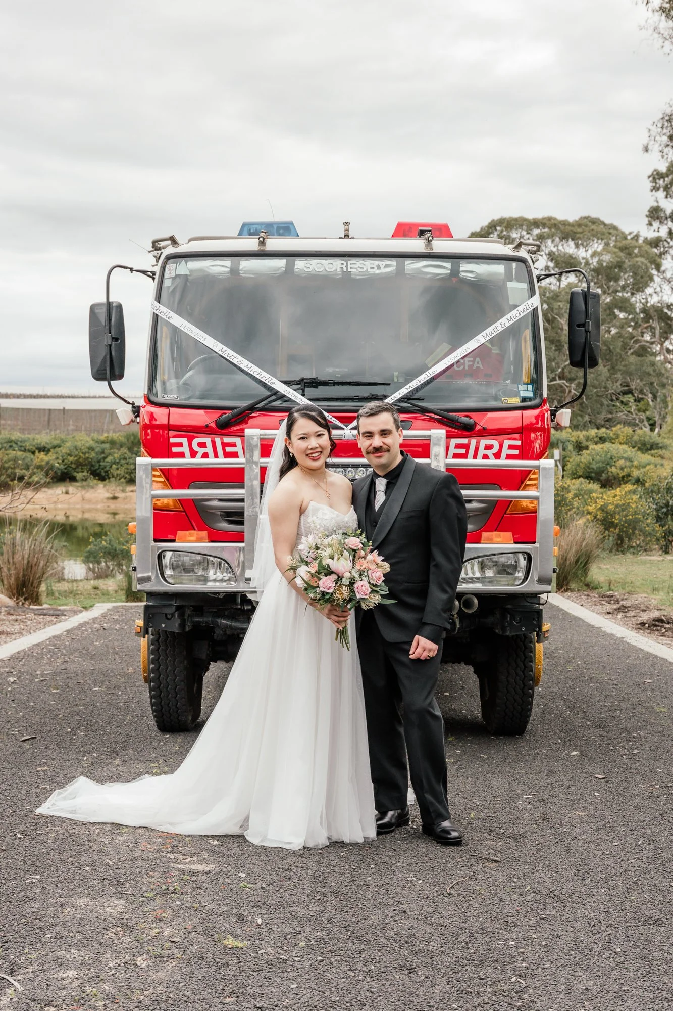 A newlywed couple standing in front of a fire truck, with the bride holding a bouquet of pink and white flowers, outdoors with trees and cloudy sky in the background.