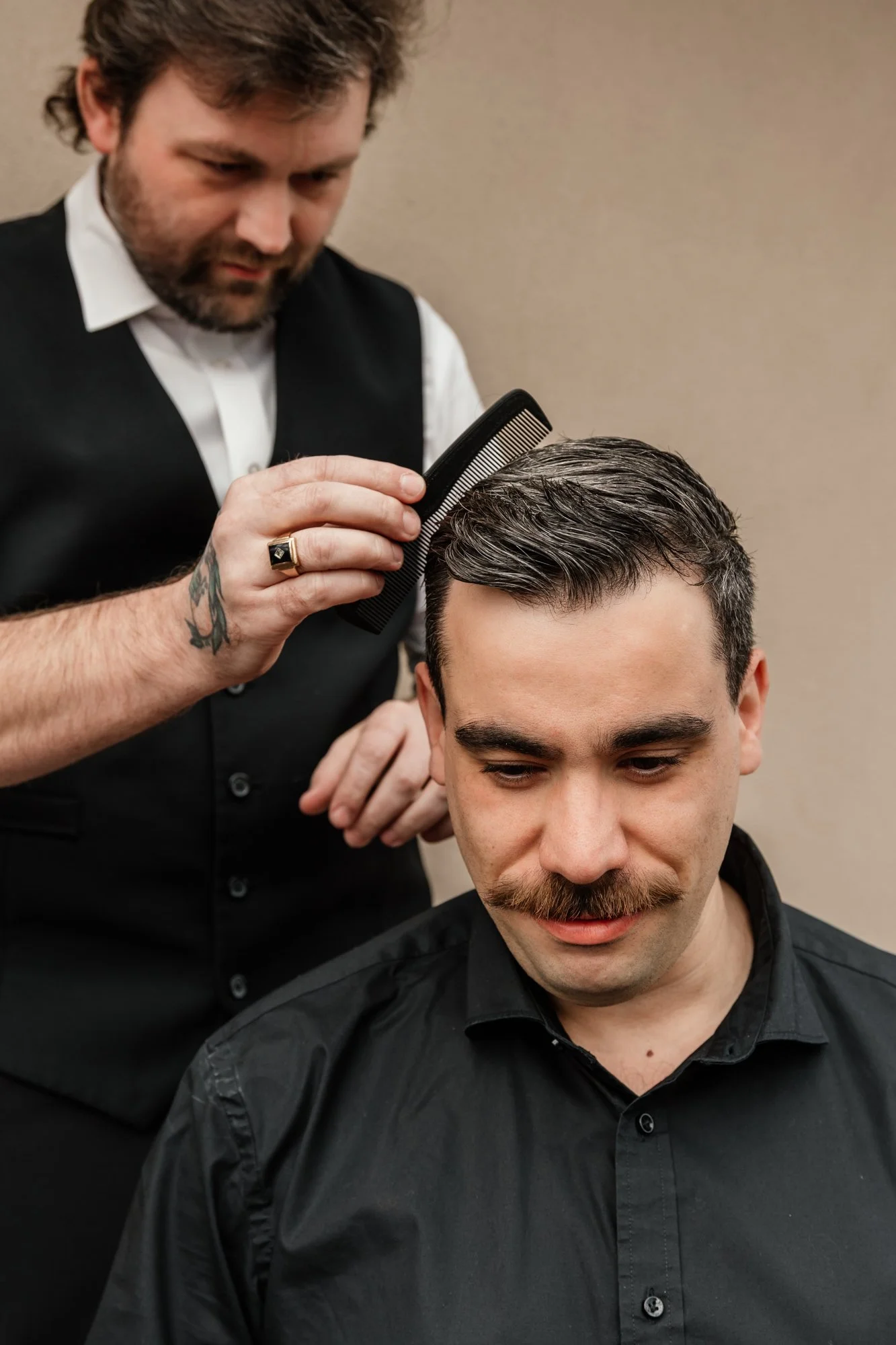 Barber combing a man's hair at a barbershop. The man has a mustache and is wearing a black shirt. The barber has tattoos on his left arm, a ring on his finger, and wears a vest over a white shirt.