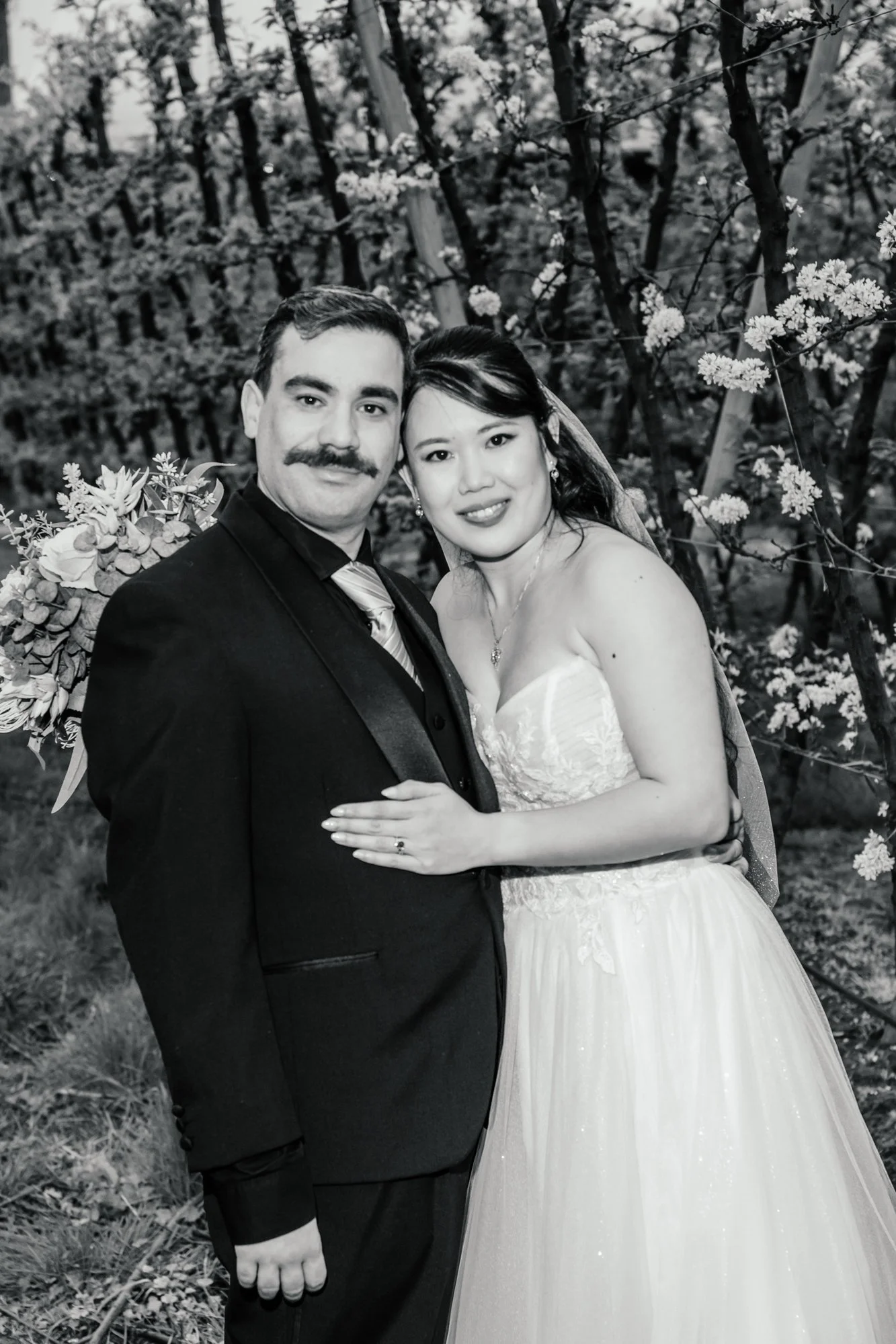 A black and white photo of a smiling couple, dressed in wedding attire, standing outdoors in front of flowering trees. The groom has a mustache and is wearing a suit, while the bride is in a strapless wedding gown with jewelry, and they are embracing