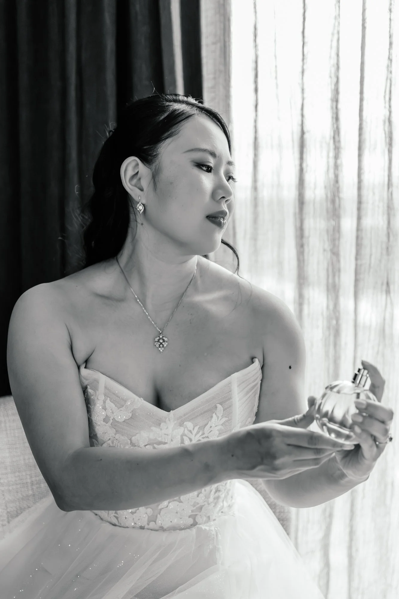 A woman in a white dress, wearing jewelry, looking at her reflection in a mirror, standing near a sheer curtain.