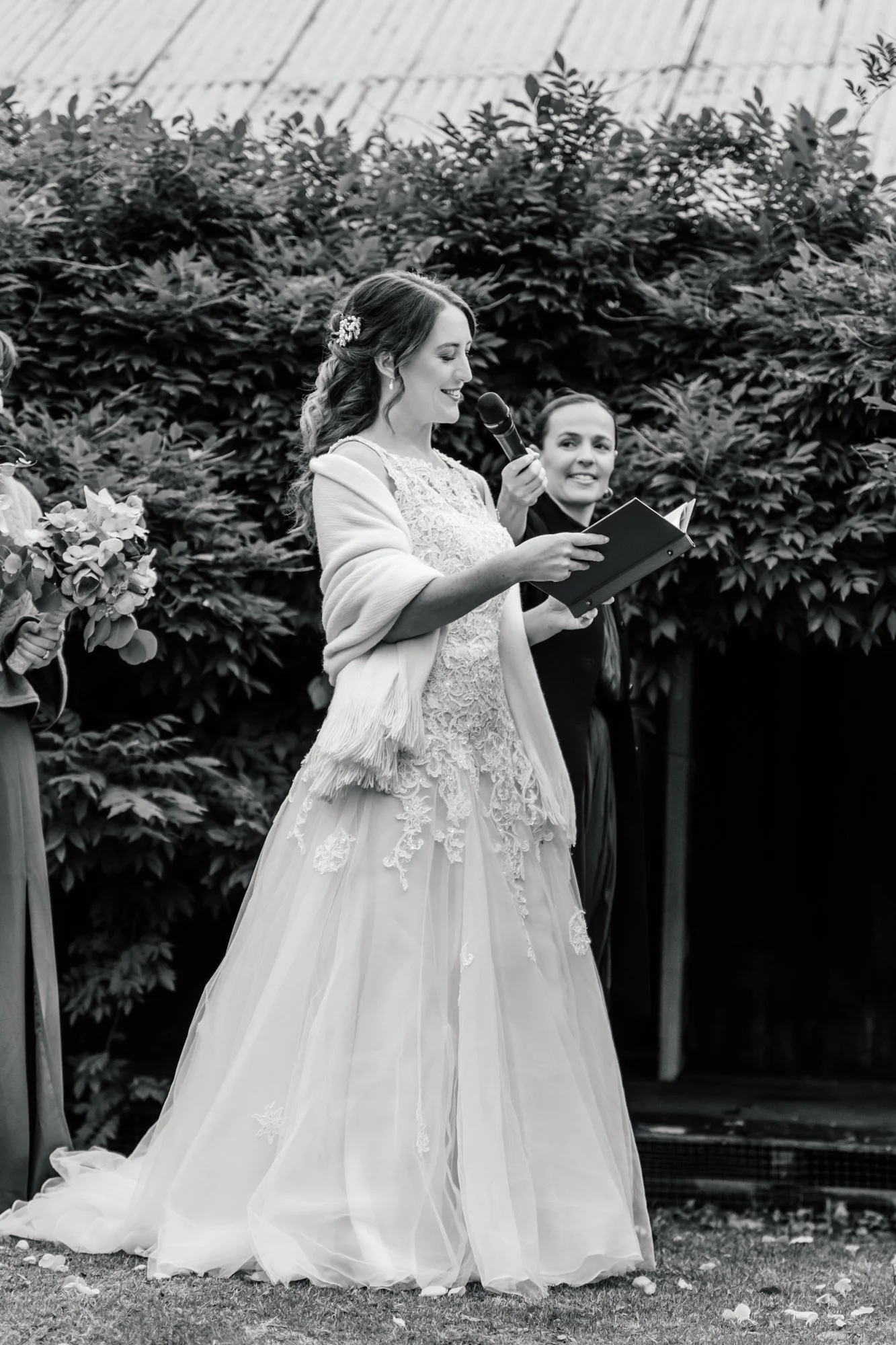 Black and white photo of a woman in a wedding dress reading from a book at an outdoor wedding ceremony.