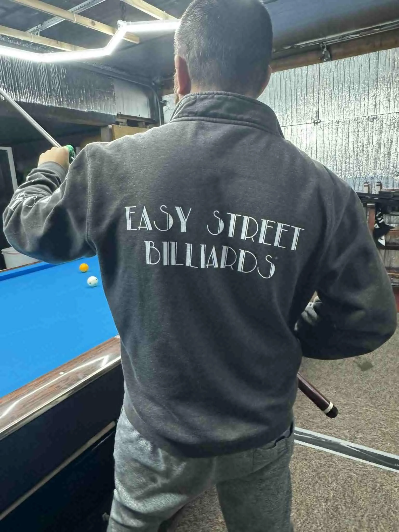 Person playing pool, wearing a 1/4-zip sweatshirt that says "Easy Street Billiards" on the back.