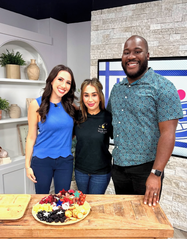 Three smiling people standing behind a wooden table with a plate of assorted berries, cookies, and festive decorations. They are indoors with a white and gray brick wall background and built-in shelves with decorative items.