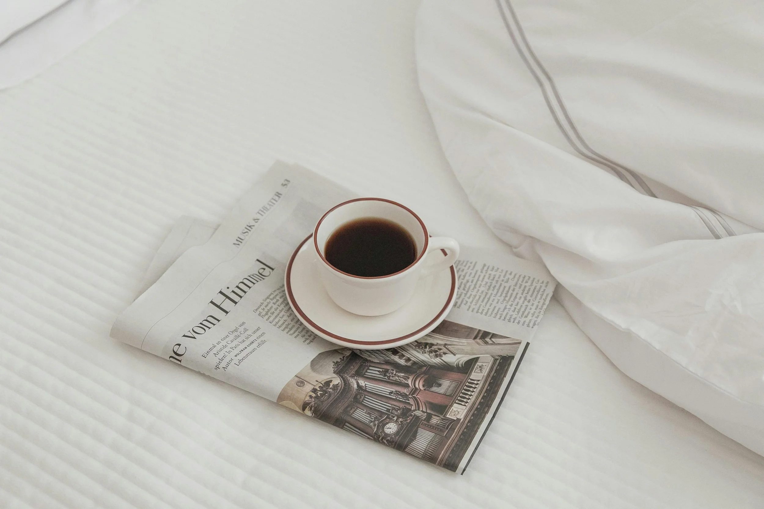 A cup of black coffee on a saucer resting on a newspaper on a white bed with white sheets and a partially visible white pillow.