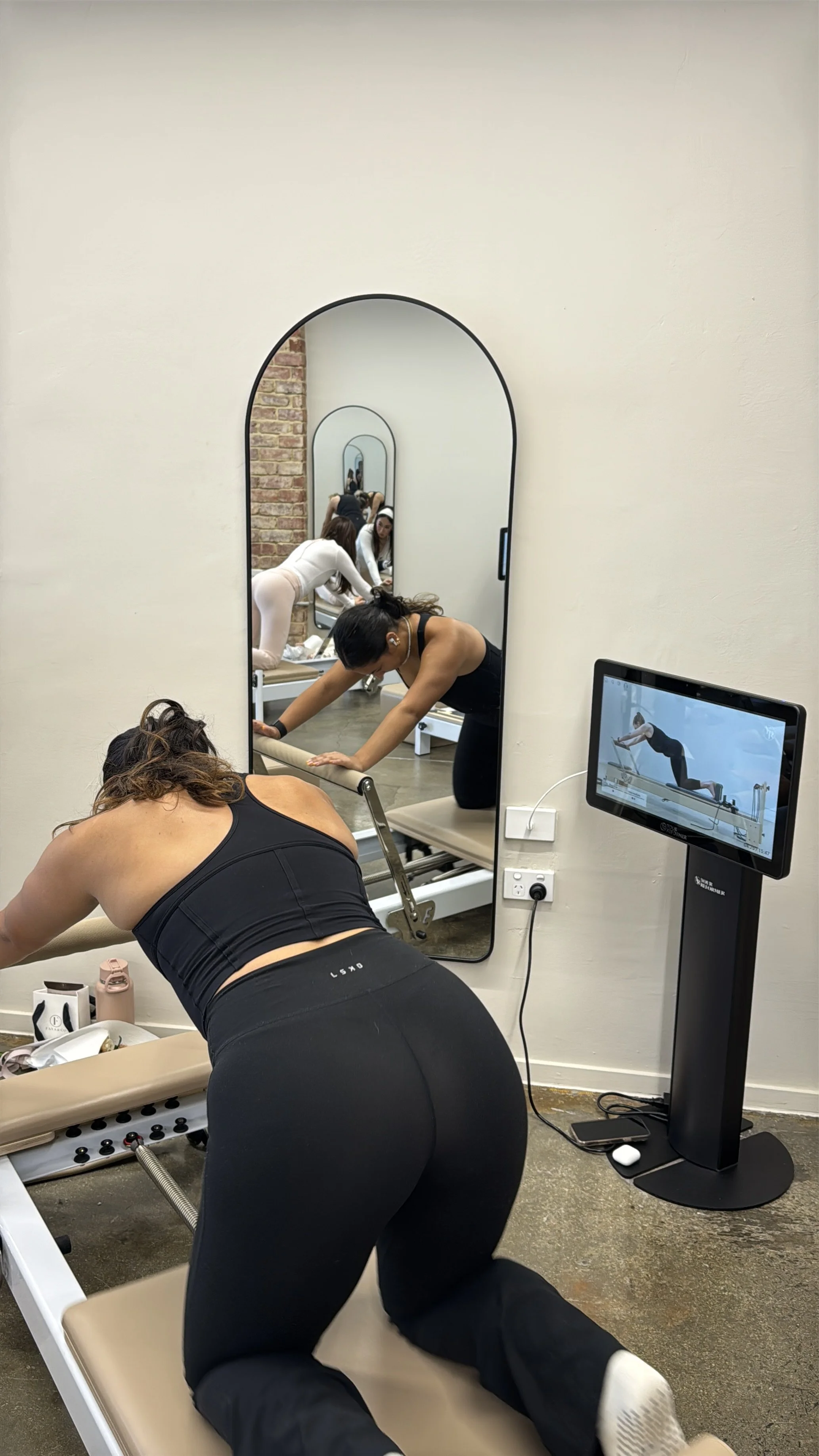 A woman doing Pilates on a reformer machine in a fitness studio with a mirror and a television monitor showing her exercise.
