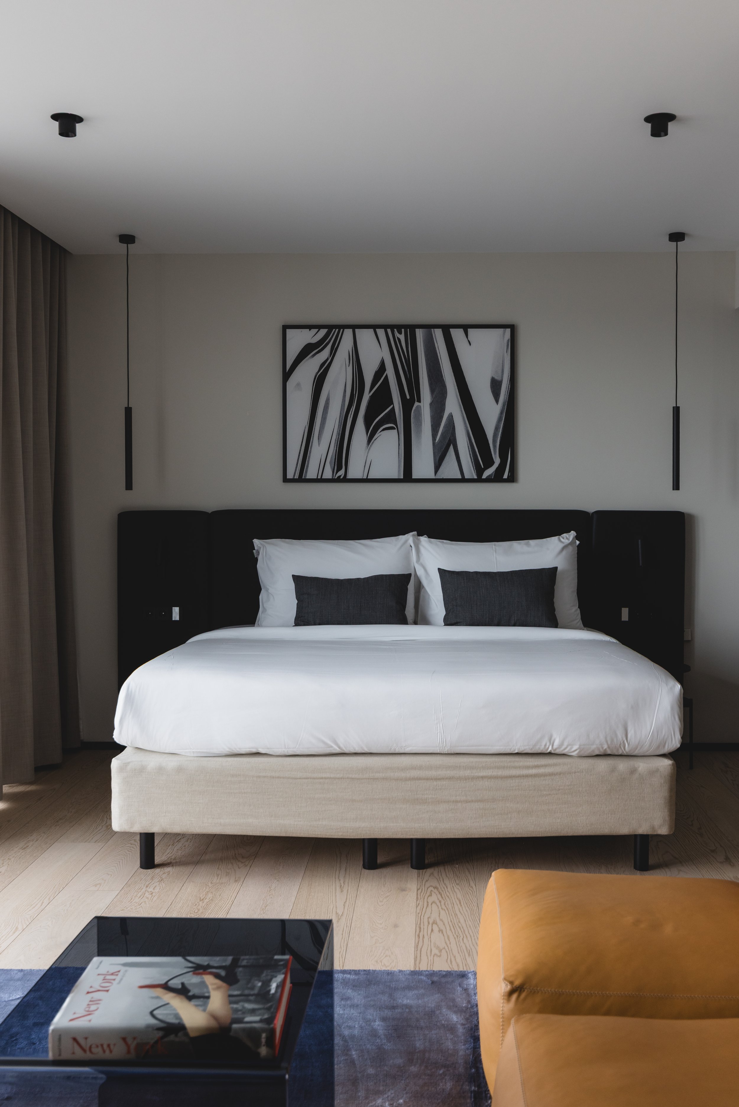 Ein Schlafzimmer mit einem großen weißen Bett, schwarzem Kopfteil und zwei weißen Kissen mit schwarzen Kissen. Über dem Bett hängt ein abstraktes schwarzes und weißes Bild. Auf dem Nachttisch liegt ein Buch. Ein brauner Sessel steht im Vordergrund.