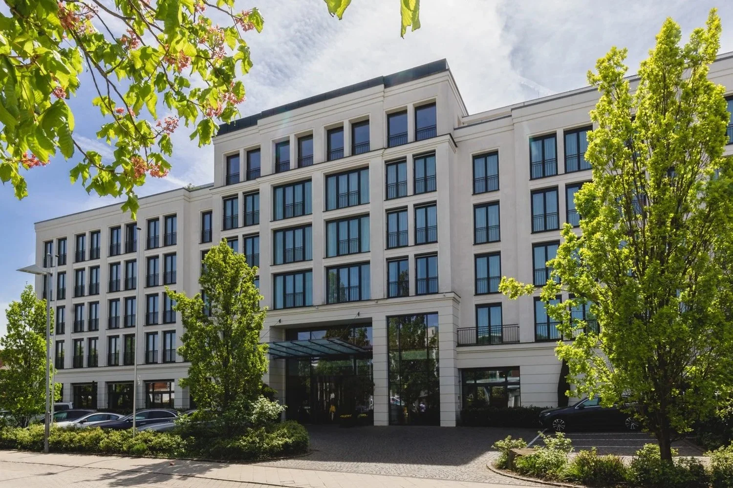 Modernes Gebäude mit mehreren Etagen und großen Fenstern, umgeben von Bäumen. Gepflasterter Vorplatz mit parkenden Autos. Sonniger Himmel und grünes Laub im Vordergrund.