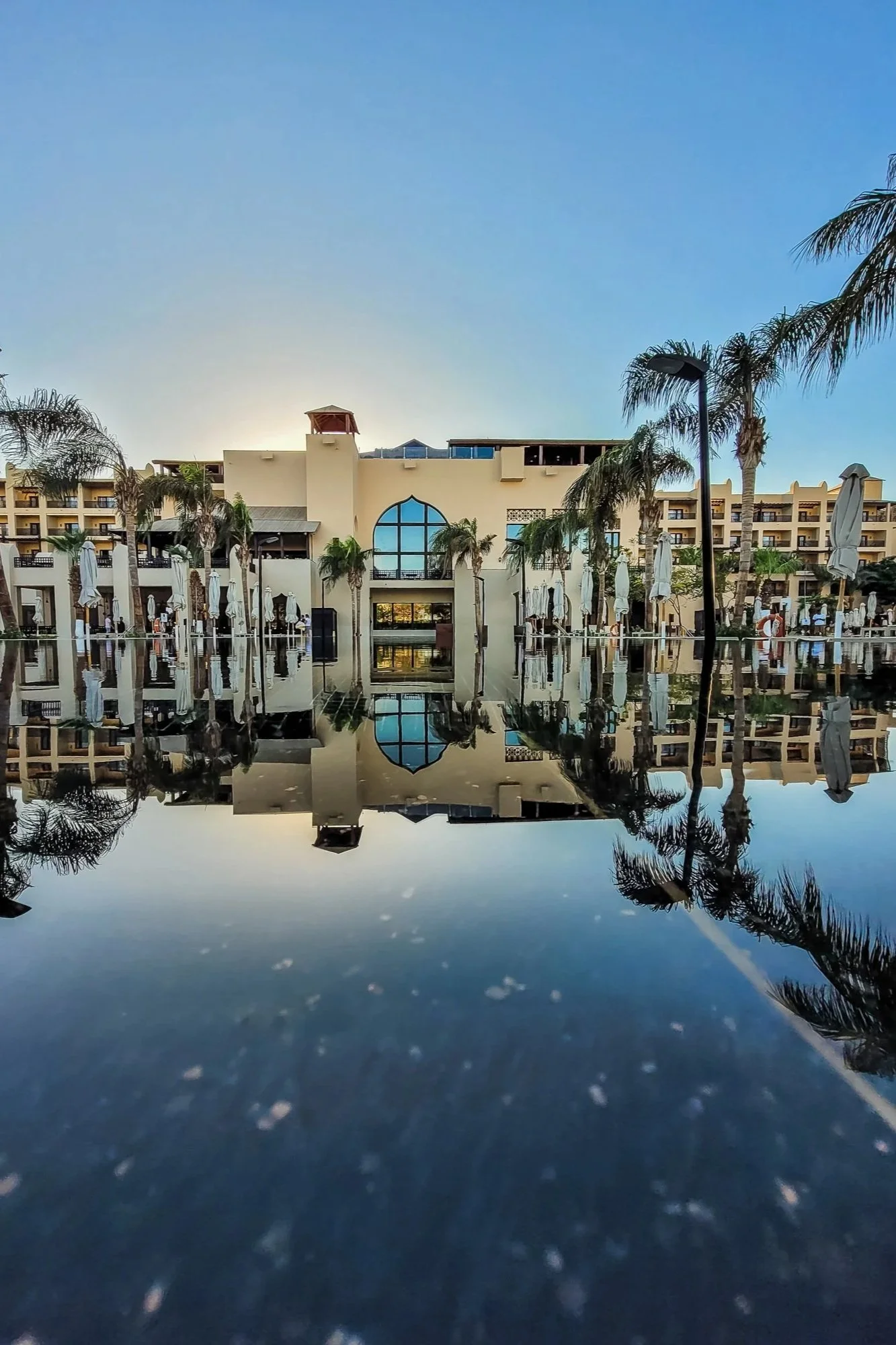 Ein Hotel mit Palmen und Sonnenschirmen, reflektiert im Wasserpool bei Sonnenuntergang.
