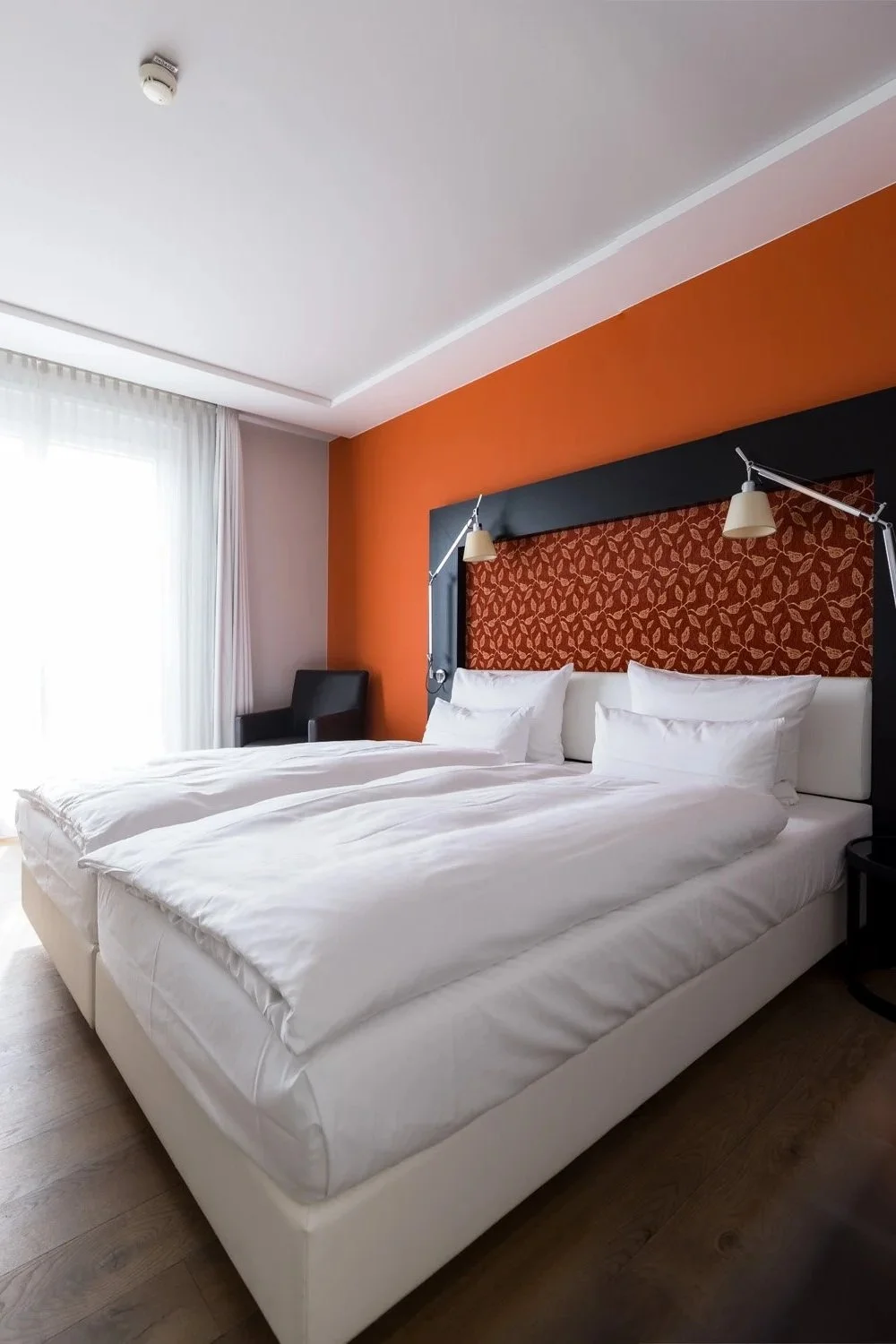 Modernes Hotelzimmer mit zwei weißen Einzelbetten, orangeroter Akzentwand, großen Fenstern mit weißen Vorhängen, Holzboden und minimalistischer Einrichtung.
