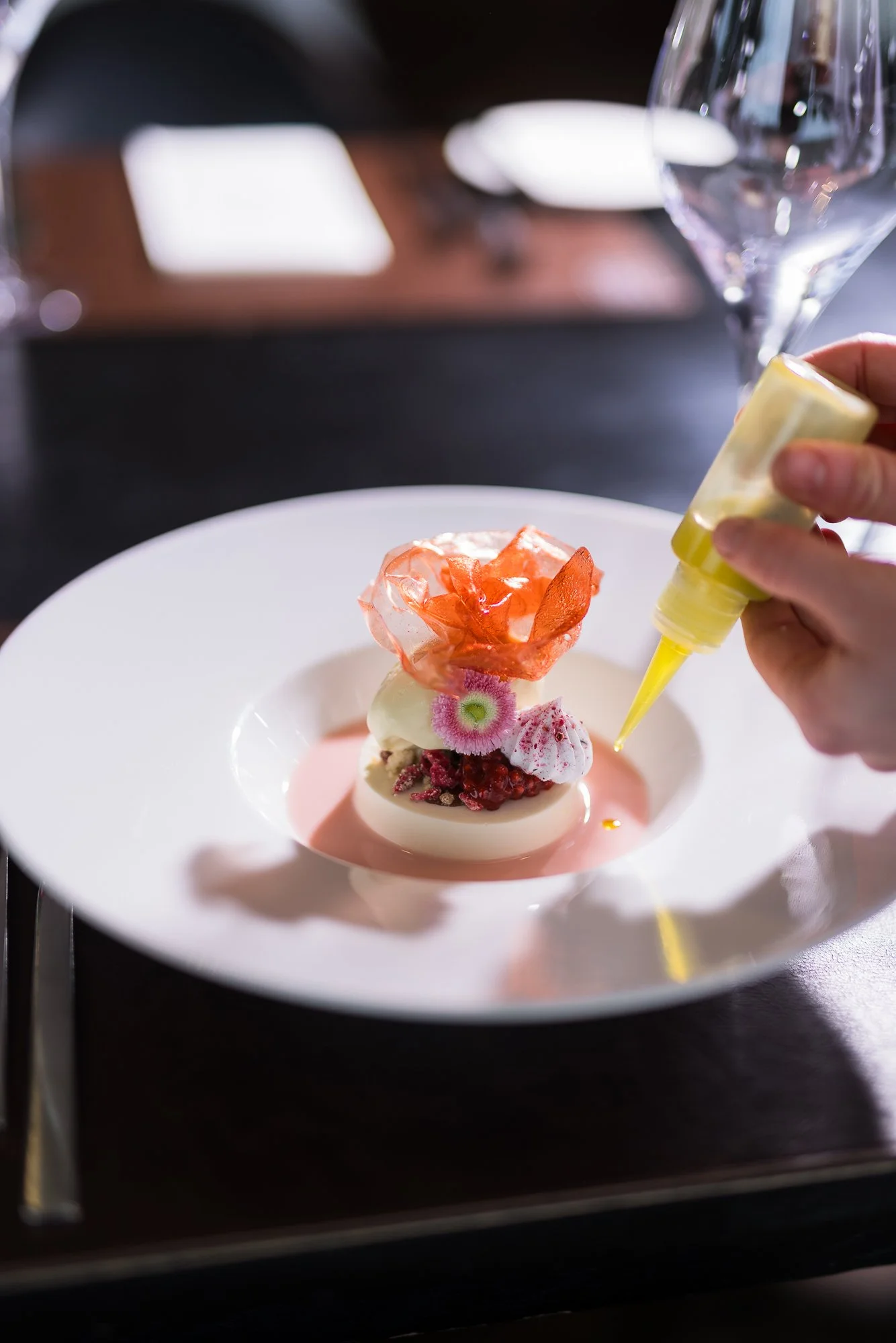 Ein Dessert auf einem weißen Teller, das mit einem Spritzsack verziert wird, inklusive Blüten und Beeren, in einer edlen Restaurant-Umgebung.