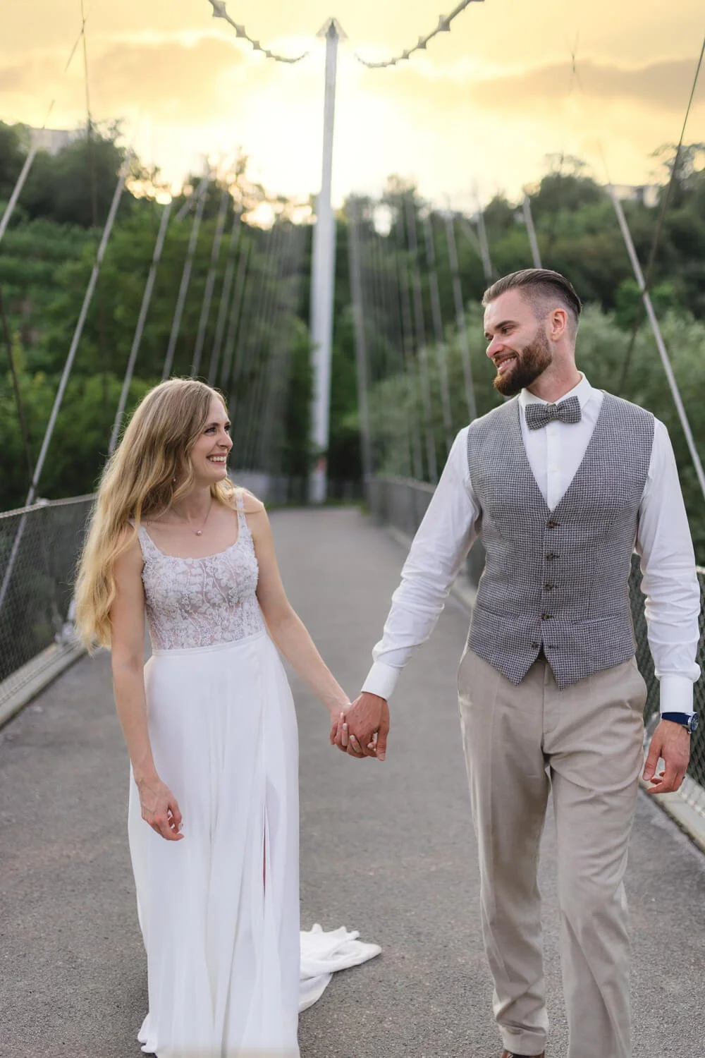 Ein Paar in eleganter Kleidung geht über eine Brücke, umgeben von Bäumen im Hintergrund.