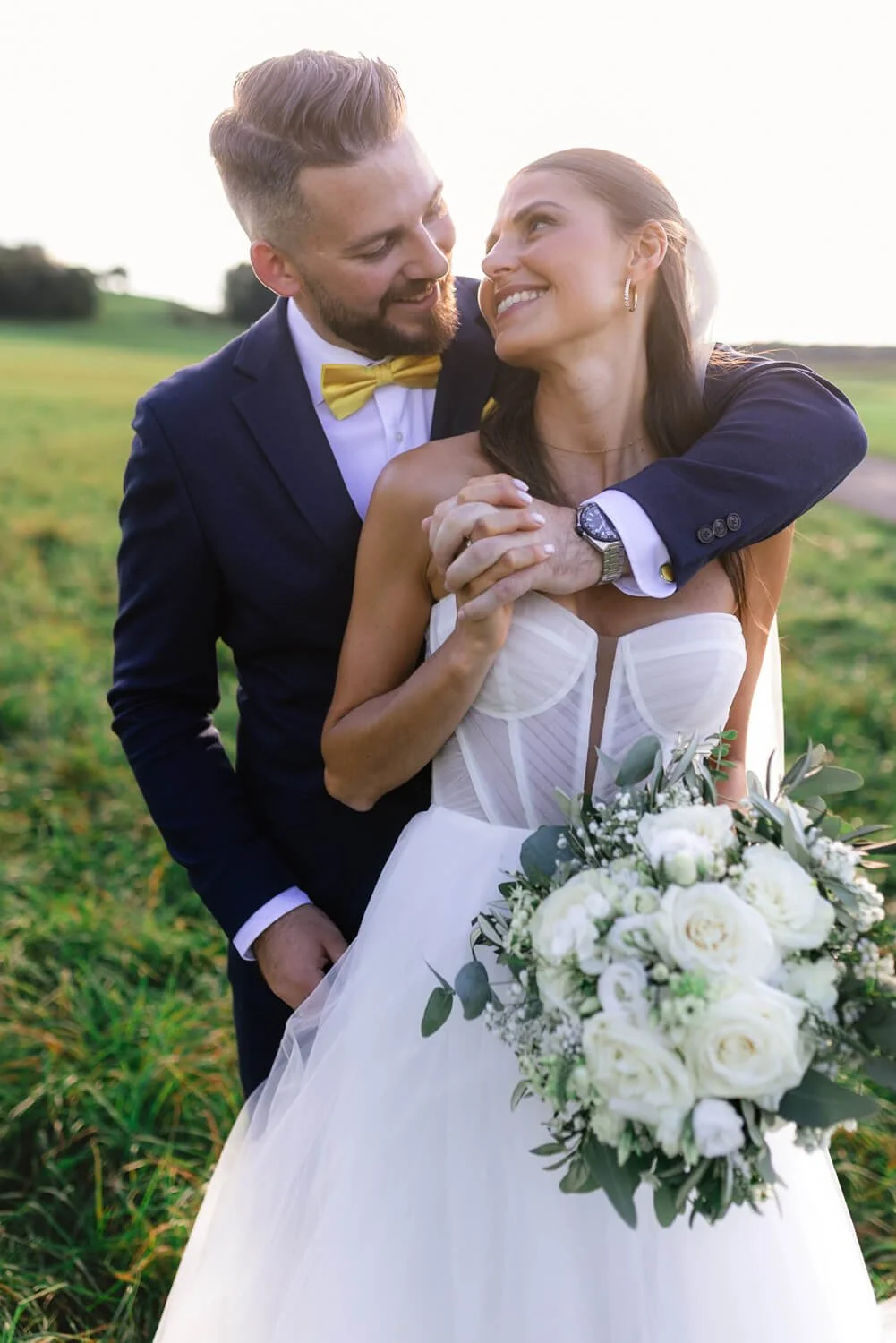 Ein Brautpaar auf einer Wiese. Die Braut trägt ein weißes Hochzeitskleid und hält einen Blumenstrauß mit weißen und grünen Blumen. Der Bräutigam trägt einen dunklen Anzug mit einer gelben Fliege. Beide lächeln und schauen sich an.