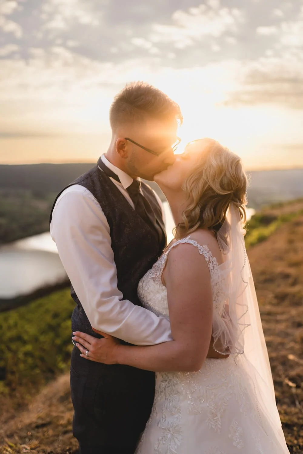 Ein Brautpaar küsst sich während ihrer Hochzeit im Freien bei Sonnenuntergang. Der Mann trägt einen Anzug, die Frau ein weißes Brautkleid mit Schleier. Im Hintergrund sind Hügel und ein Fluss zu sehen.
