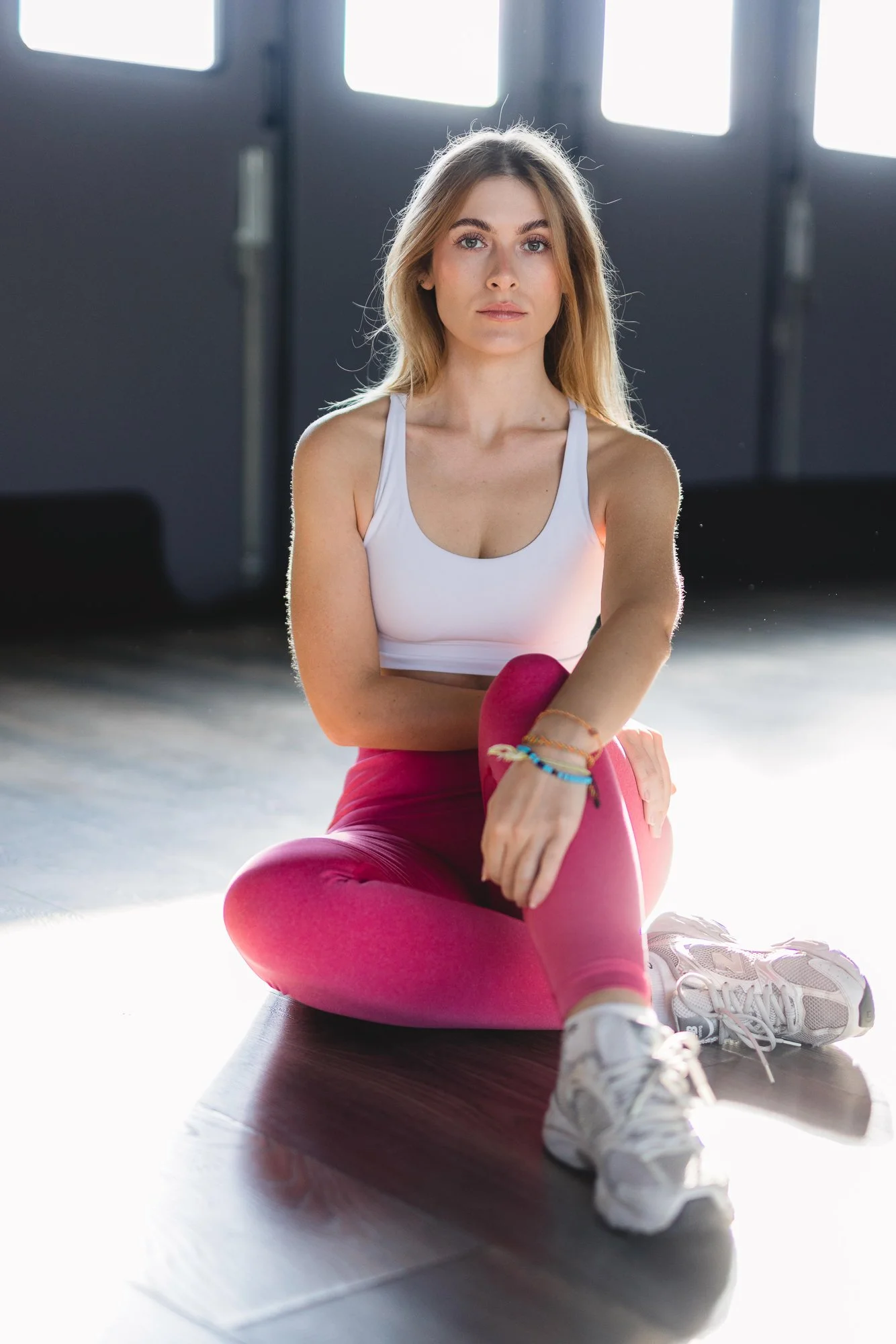 Junge Frau, sportlich, trägt weißen Sportswear und pinke Leggings, sitzt auf einer Yoga-Matte im Fitnessstudio, mit Sonnenlicht im Hintergrund.