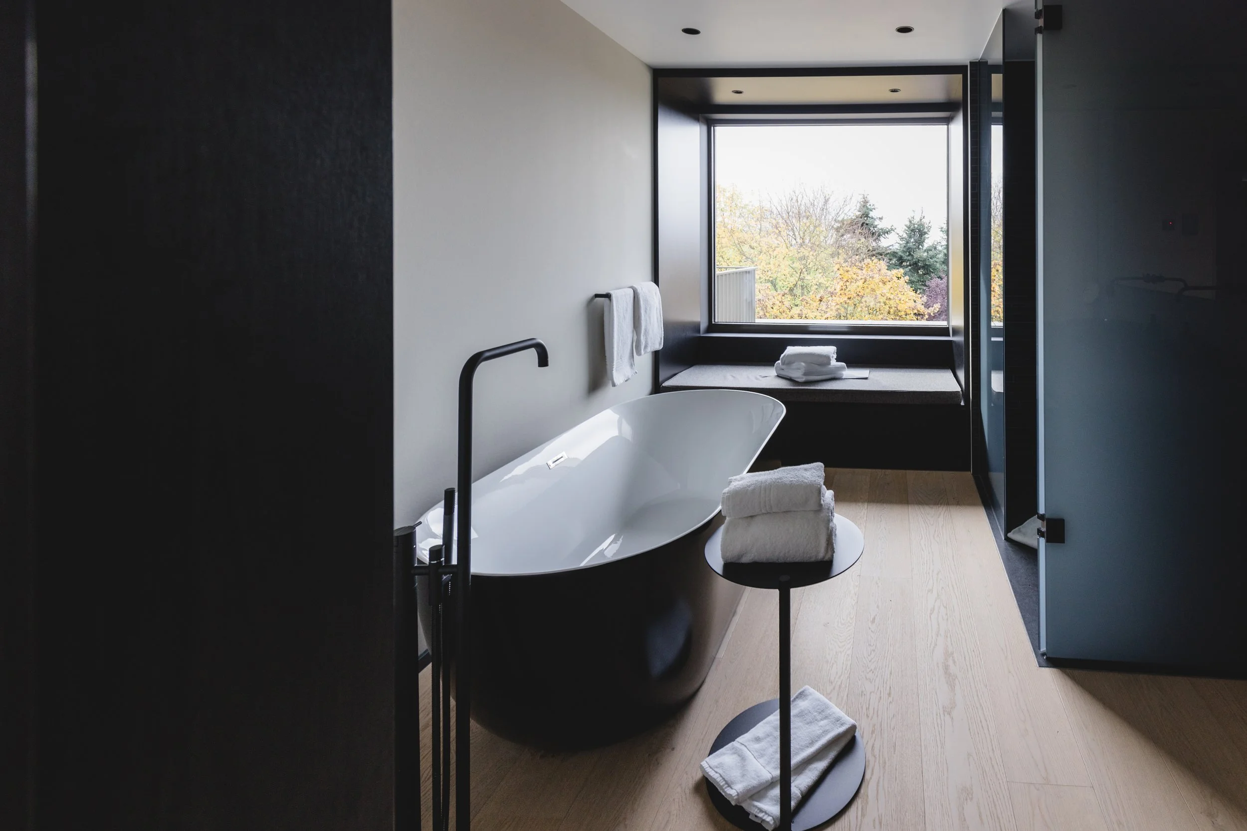 Ein modernes Badezimmer mit freistehender Badewanne, einem Fenster mit Blick auf Bäume, Handtüchern auf einem kleinen Tisch, und einem Sitzbereich im Fensterraum.