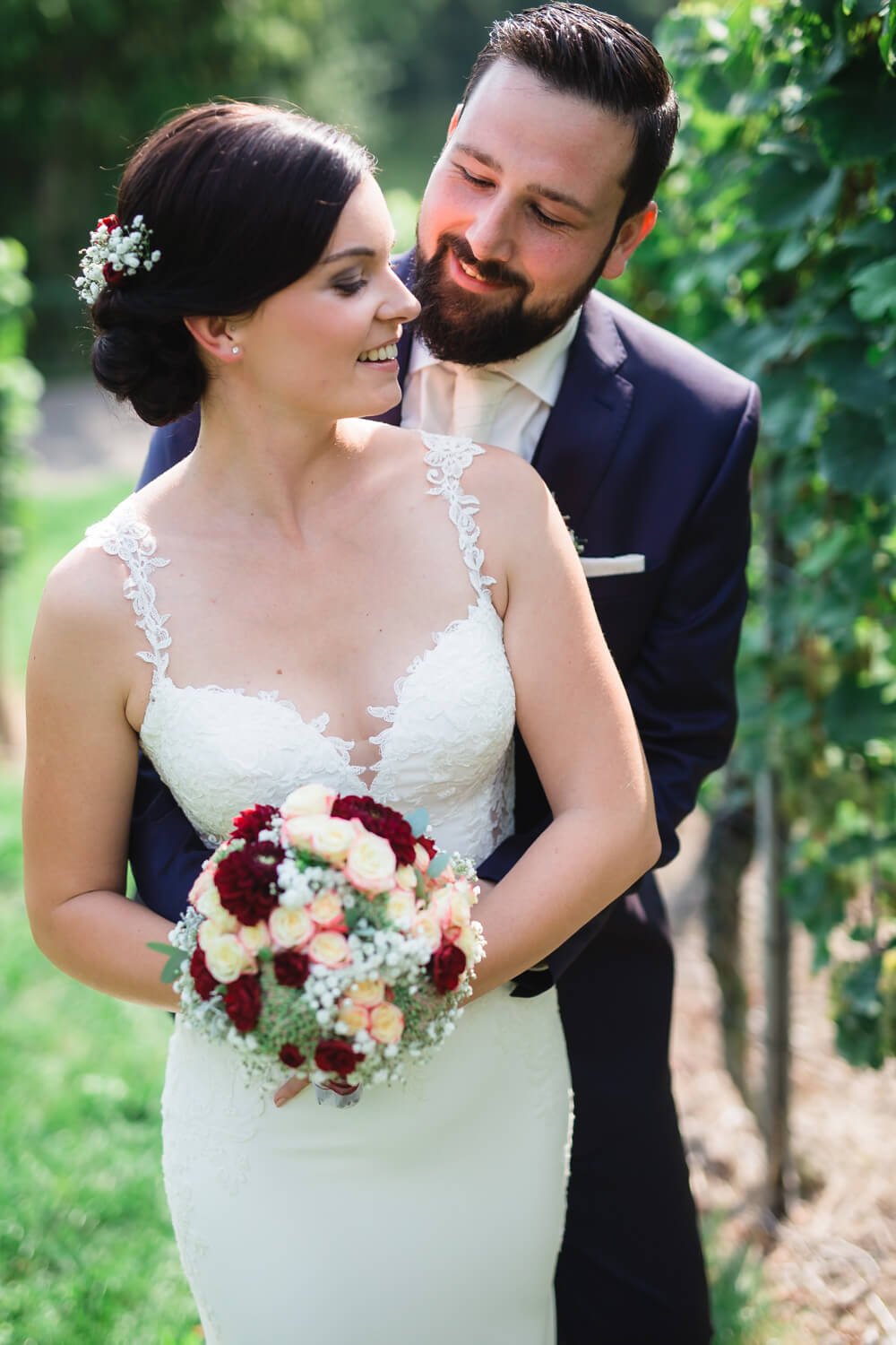 Ein Hochzeitspaar in einem Weinberg, die Braut in einem weißen Kleid mit Blumenstrauß, der Bräutigam im Anzug.