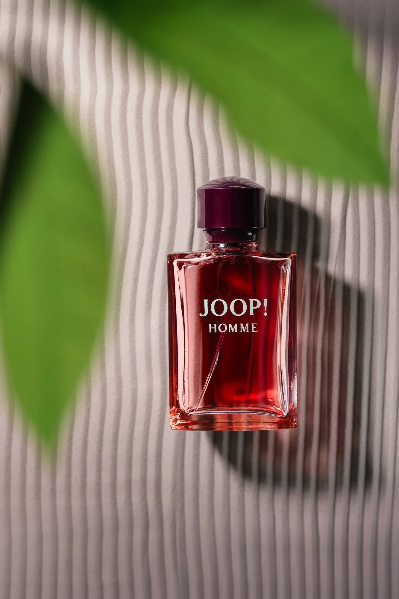 Ein rotes Fläschchen von Joop! Homme auf einer gestreiften, grauen Oberfläche, teilweise verdeckt durch grüne Blätter im Vordergrund.