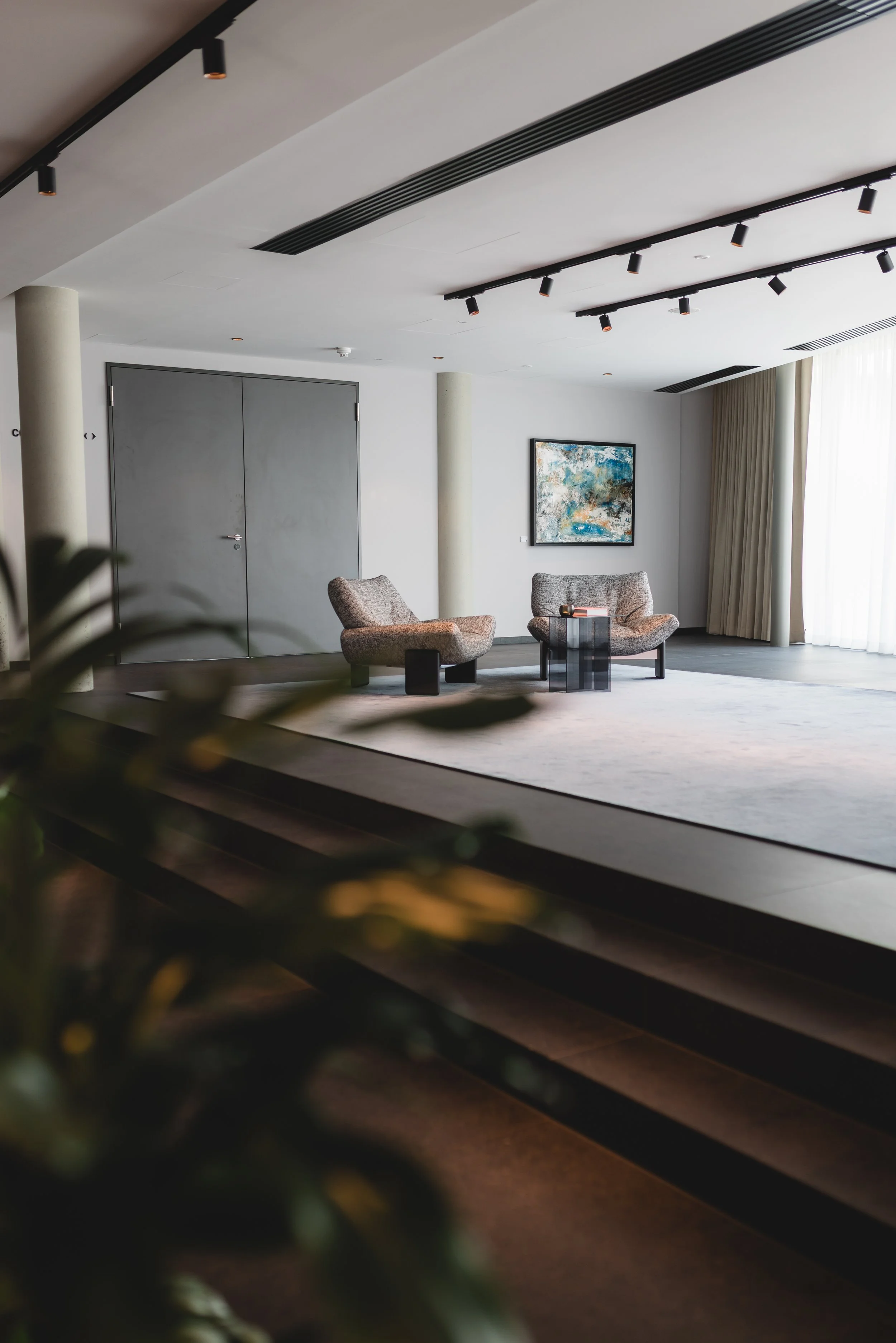 Moderner Lounge-Bereich mit zwei bequemen Sessel, einem kleinen Tisch, einem abstrakten Gemälde an der Wand und großen Fenstern mit Vorhängen.