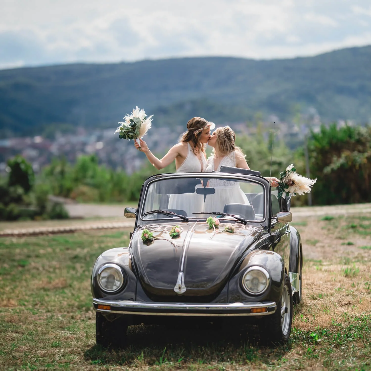 Zwei Frauen in weißen Kleidern, die sich küssen, stehen in einem offenen, dekorierten Vintage-Auto im Freien mit einem malerischen Hintergrund aus Hügeln und Wolken.