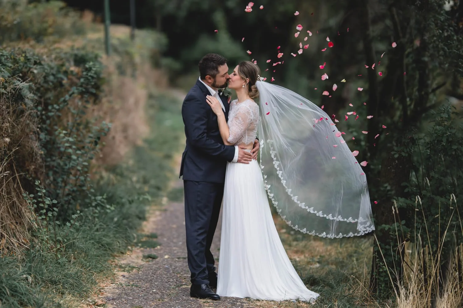 Ein Brautpaar küsst sich im Freien, umgeben von fallenden Blütenblättern. Die Braut trägt ein langes weißes Kleid und einen Schleier. Der Bräutigam ist in einem dunklen Anzug gekleidet. Im Hintergrund ist grüne Vegetation zu sehen.