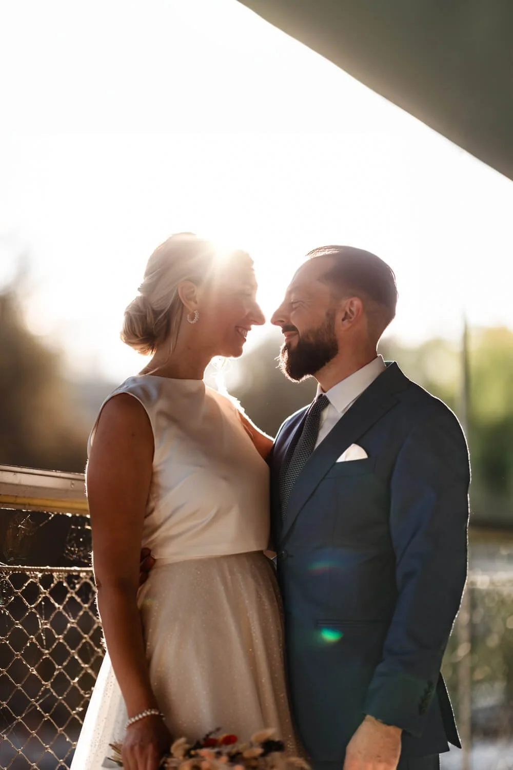 Ein Brautpaar steht einander zugewandt in der Sonne, die Braut trägt ein weißes Kleid und der Bräutigam einen dunklen Anzug.