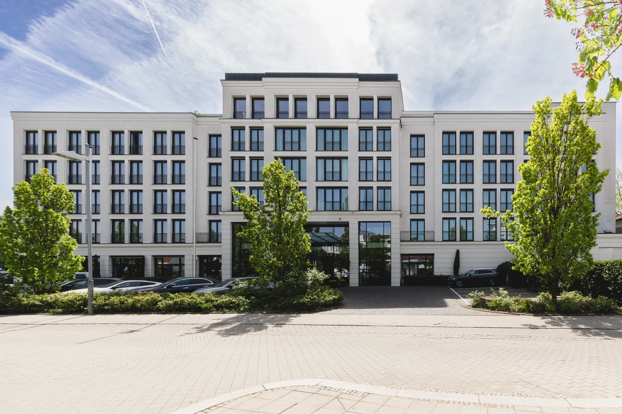 Moderner weiße Hotel mit mehreren Fenstern, umgeben von grünen Bäumen und Parkplätzen, bei Tageslicht und bewölktem Himmel.