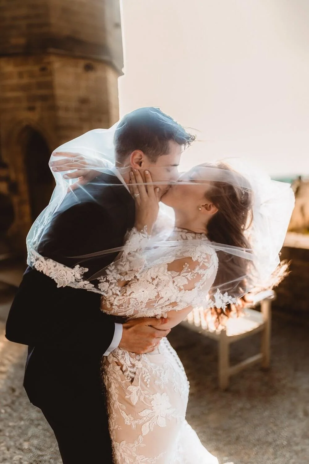 Braut und Bräutigam küssen sich unter einem Schleier bei einem Fotoshooting. Die Braut trägt ein Spitzenkleid, der Bräutigam einen Anzug. Romantische Hochzeitsatmosphäre.