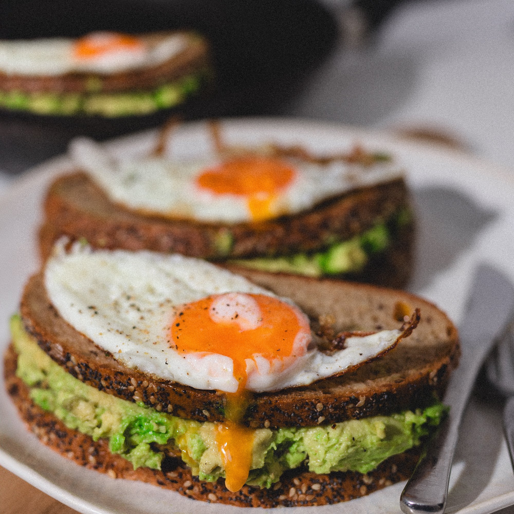 Blick auf eine gekochte Eierhälfte mit flüssigem Eigelb, auf einem Teller mit Avocado, Tomate und dunklem Brot. Ein Messer liegt am Teller.