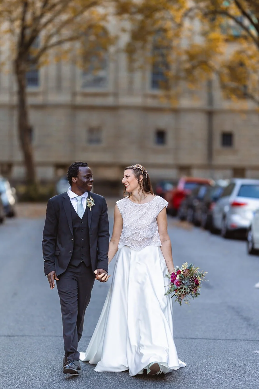 Ein Brautpaar in Hochzeitskleidung geht Hand in Hand auf einer Straße. Die Braut trägt ein weißes Kleid und hält einen Blumenstrauß, während der Bräutigam einen dunklen Anzug trägt. Im Hintergrund sind unscharf Bäume und Gebäude zu sehen.
