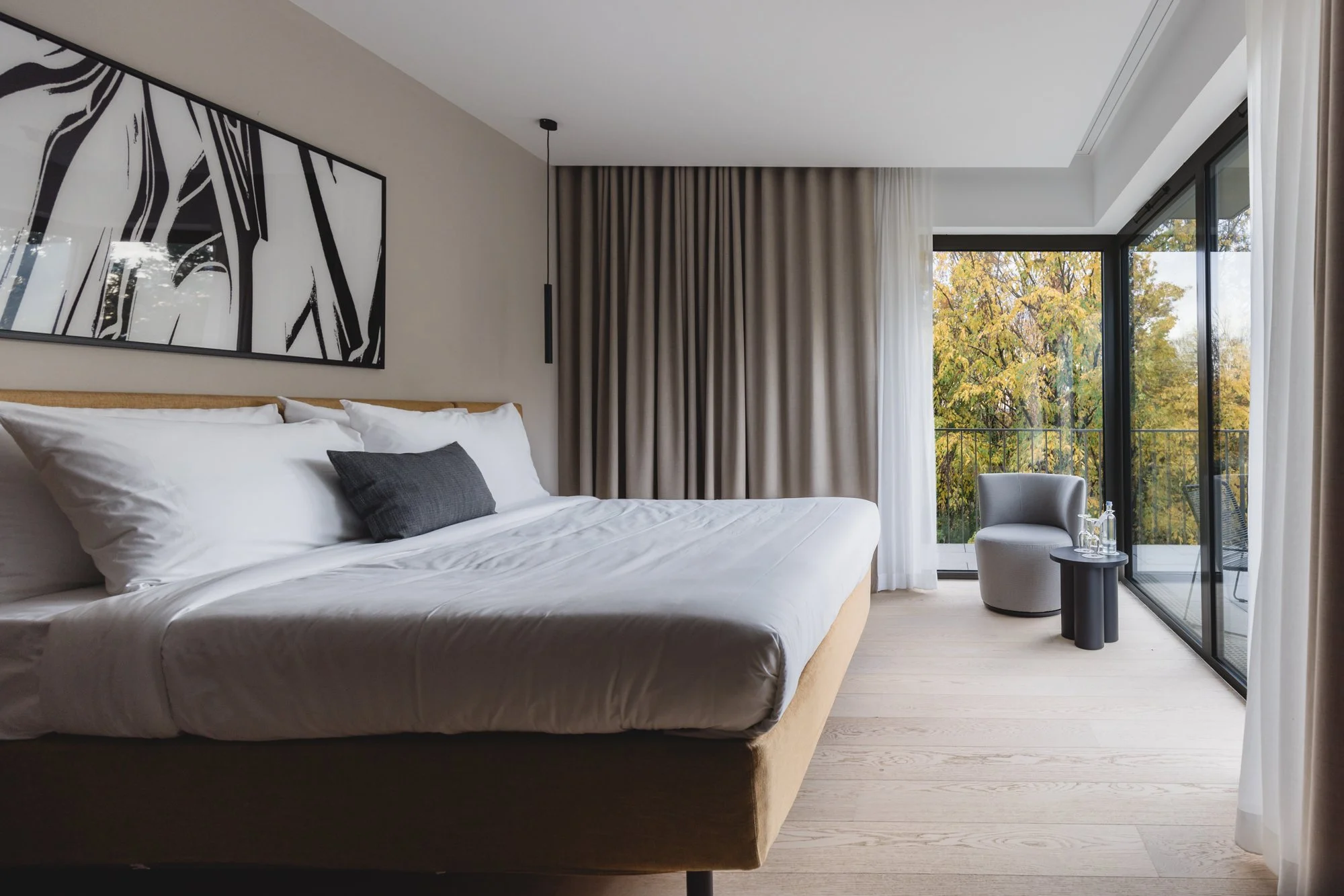 Schlafzimmer mit großem Bett, Wandkunst, Vorhängen, Sessel, kleiner Beistelltisch mit Wasserflaschen, Balkon mit Glasfront und Blick auf herbstliche Bäume.