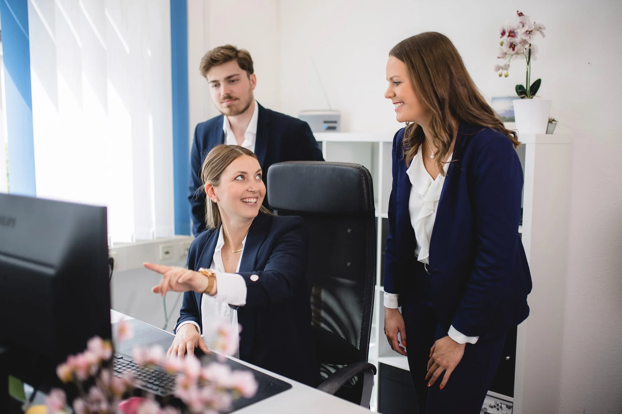 Zwei Geschäftsfrauen im Büro, wobei eine Frau an einem Computer sitzt und mit einer anderen spricht, die lächelnd steht, während im Hintergrund ein Mann zuschaut.