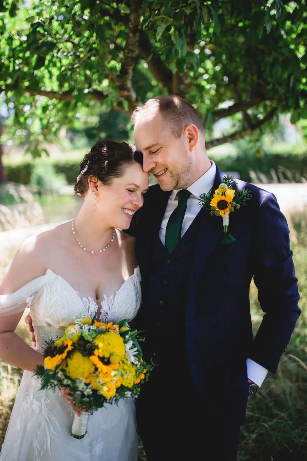 Brautpaar lächelnd unter einem Baum, Braut in weißem Kleid mit Blumenstrauß, Bräutigam im dunkelblauen Anzug mit Sonnenblumenanstecker, sonniger Tag.