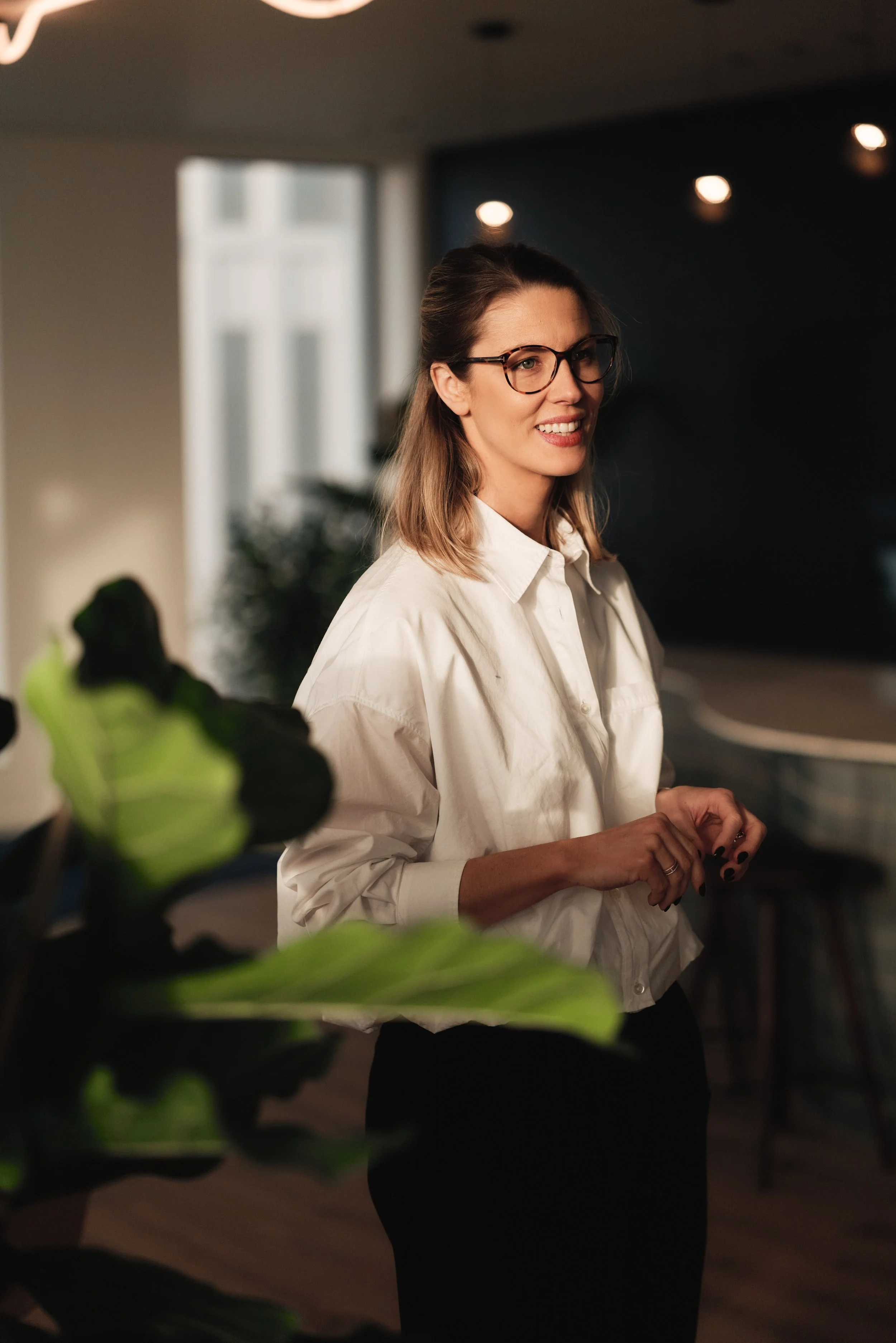 Vrouw met bruin haar, brillen en witte blouse in een interieur met planten en donkere achtergrond.