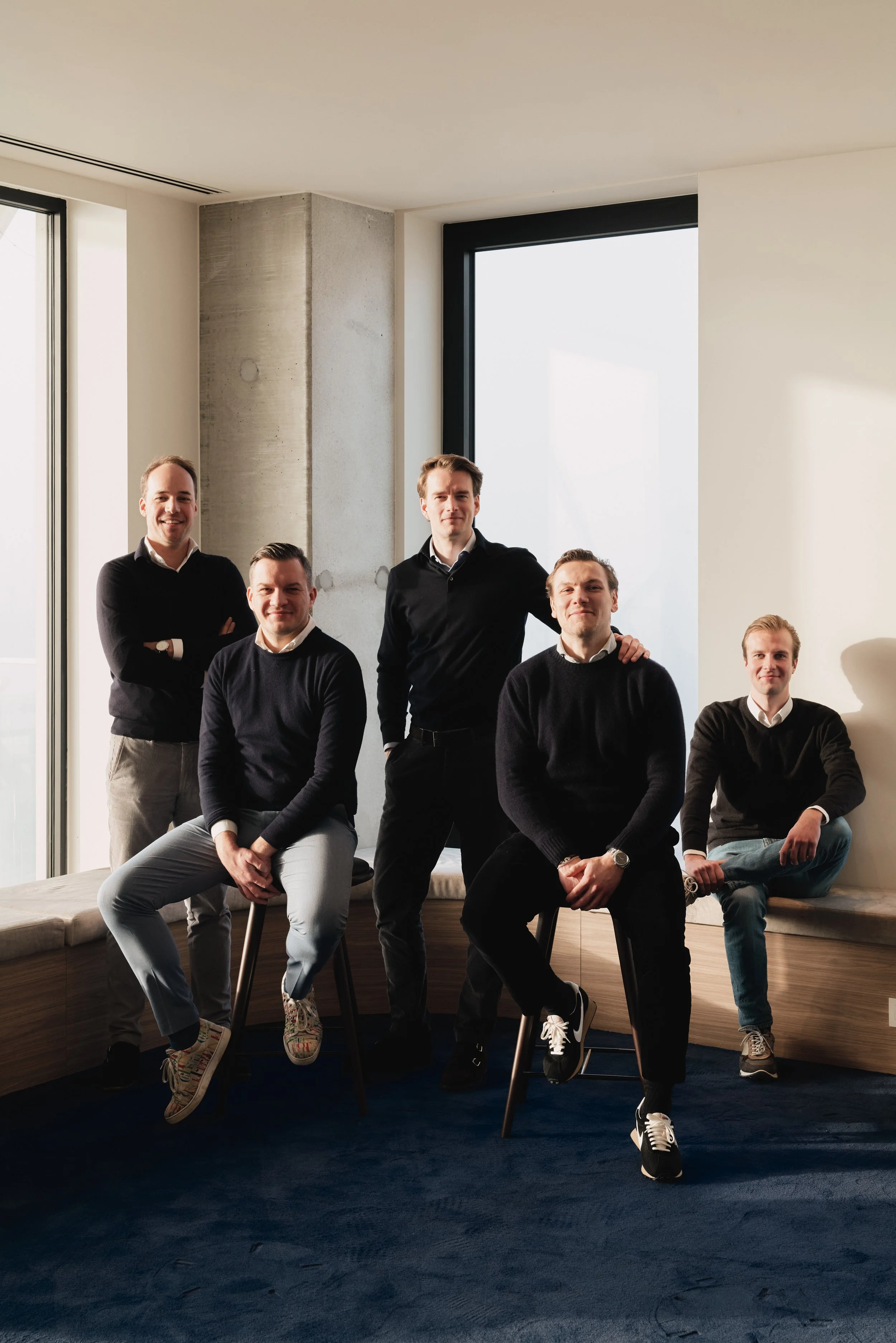 Zes mannen poseren in een modern, minimalistisch interieur met grote ramen en een blauwe vloer.