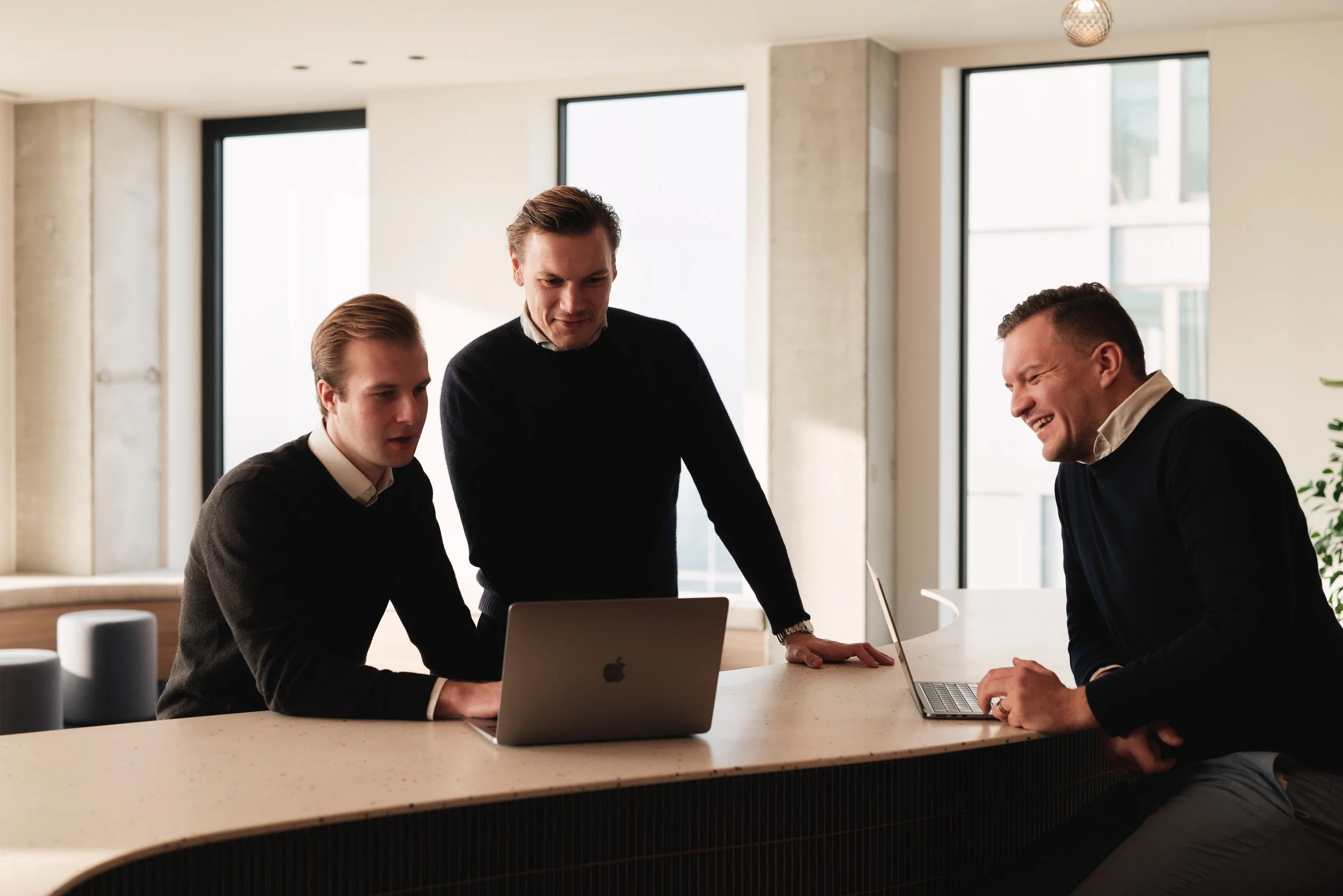 Drie zakenmensen praten en lachen samen in een vergaderzaal met grote ramen, terwijl ze werken met laptops.