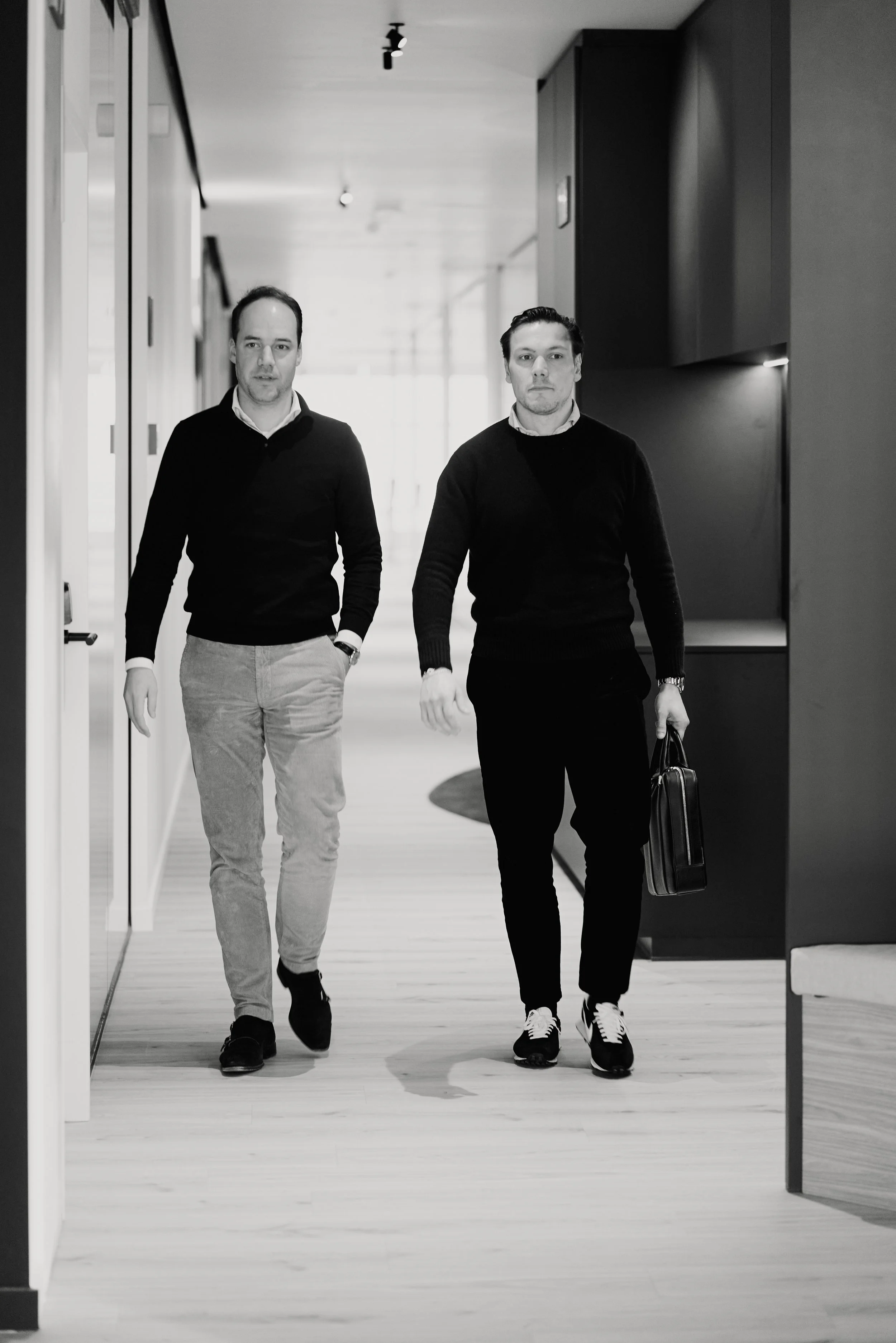 Twee jonge mannen wandelen door een modern, kleurrijk lobbygebied, gekleed in casual zakelijke kleding, een draagt een tas.