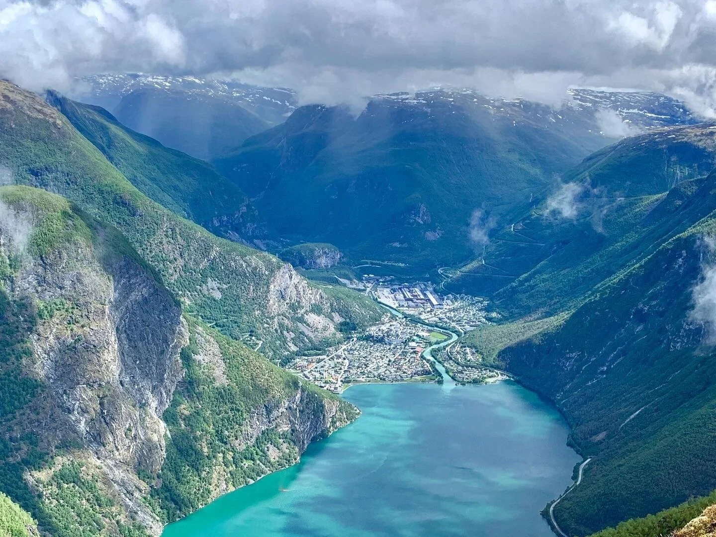 Fjell og innsjøer i en norsk fjord med en liten by langs vannkanten, omgitt av grønne åser og høye fjell med noe snø på toppene.