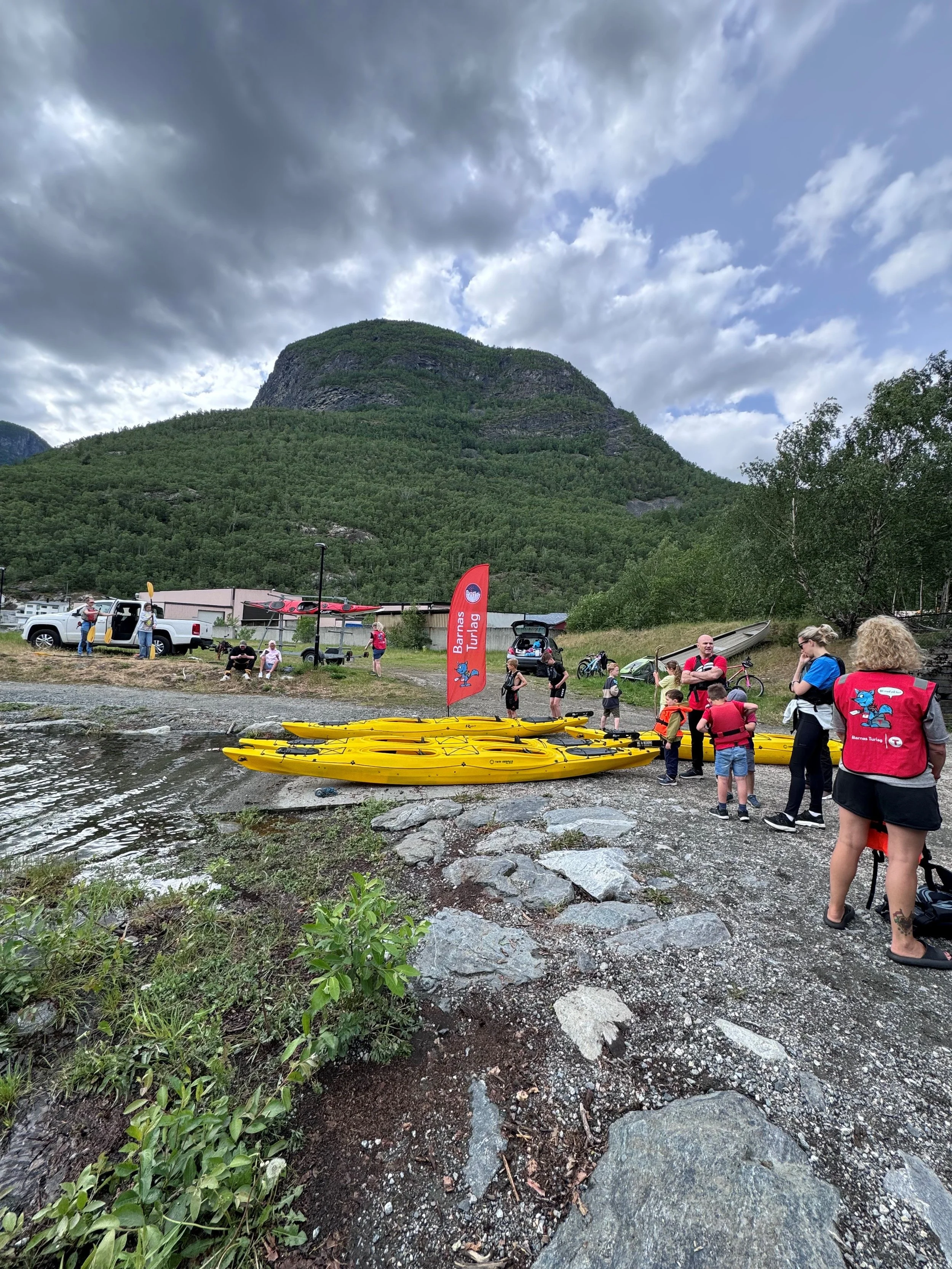 Folk samles ved vannkanten med utstyr for kajakkpadling, omgitt av en fjellrik natur. Det er flere mennesker, noen med redningsvester, og flere gule kajakker ligger på bakken.