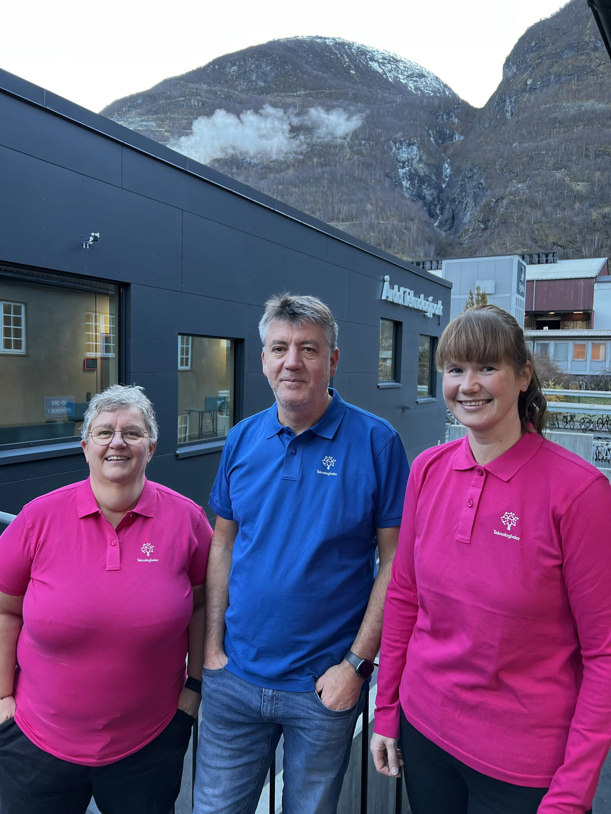 Tre mennesker foran en bygning med fjell i bakgrunnen, to kvinner i rosa og en mann i blått.