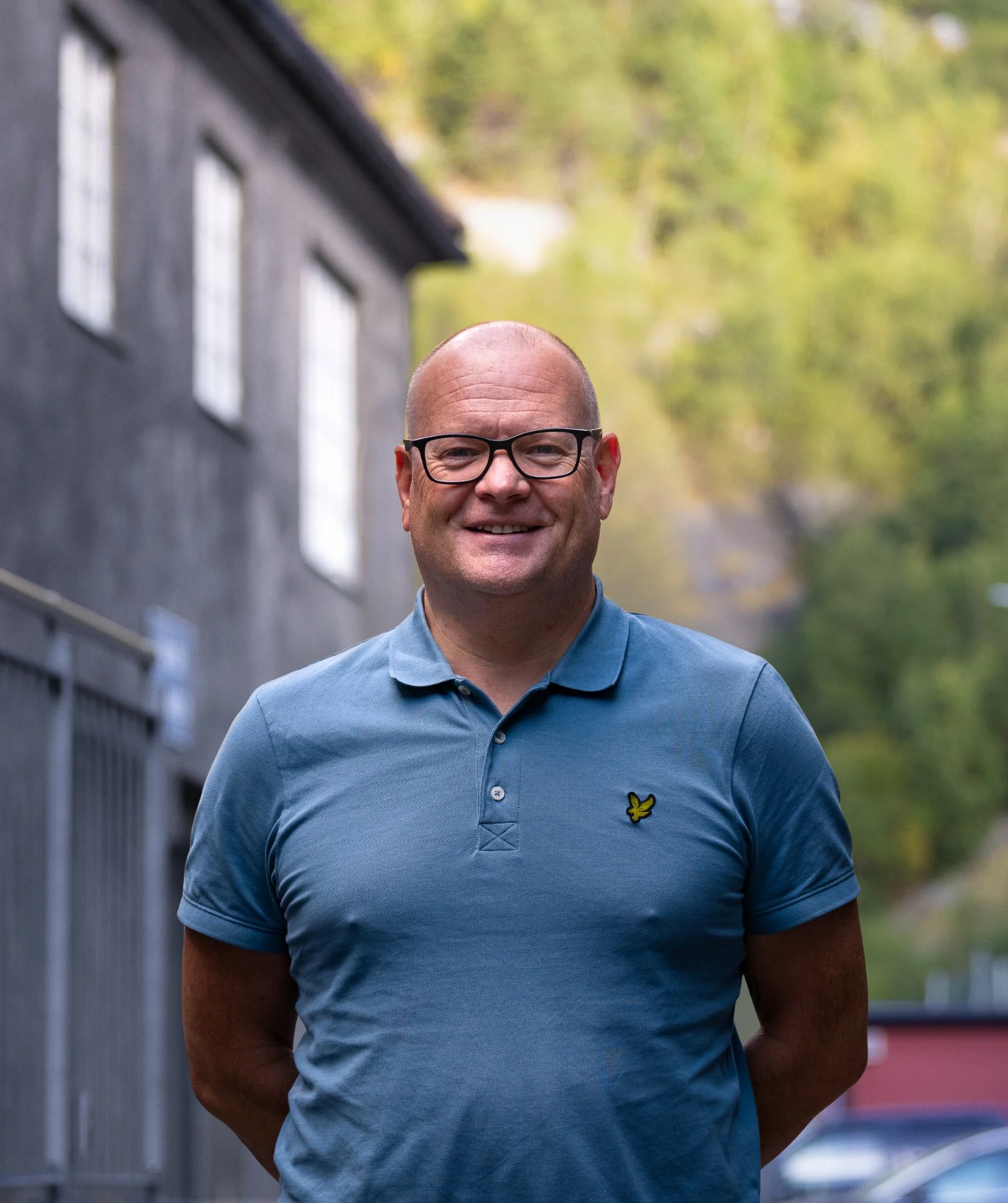 En smilende, bermen mann med briller i en blå polo trøye står utendørs, med et hus vegg og grønne trær i bakgrunnen.