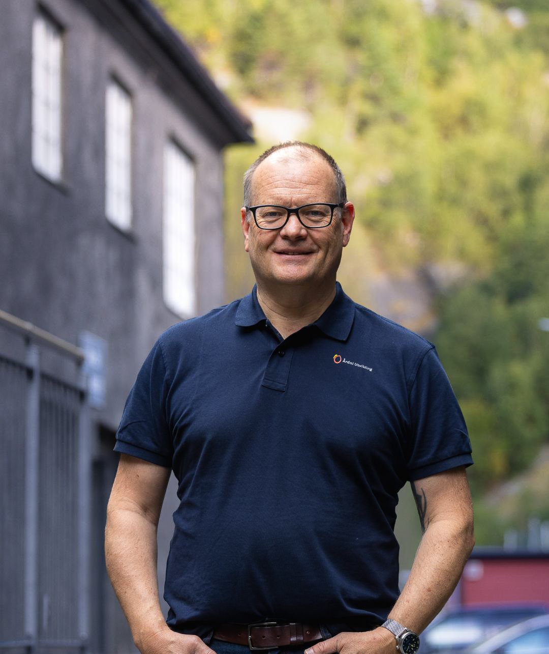 En smilende, bermen mann med briller i en blå polo trøye står utendørs, med et hus vegg og grønne trær i bakgrunnen.