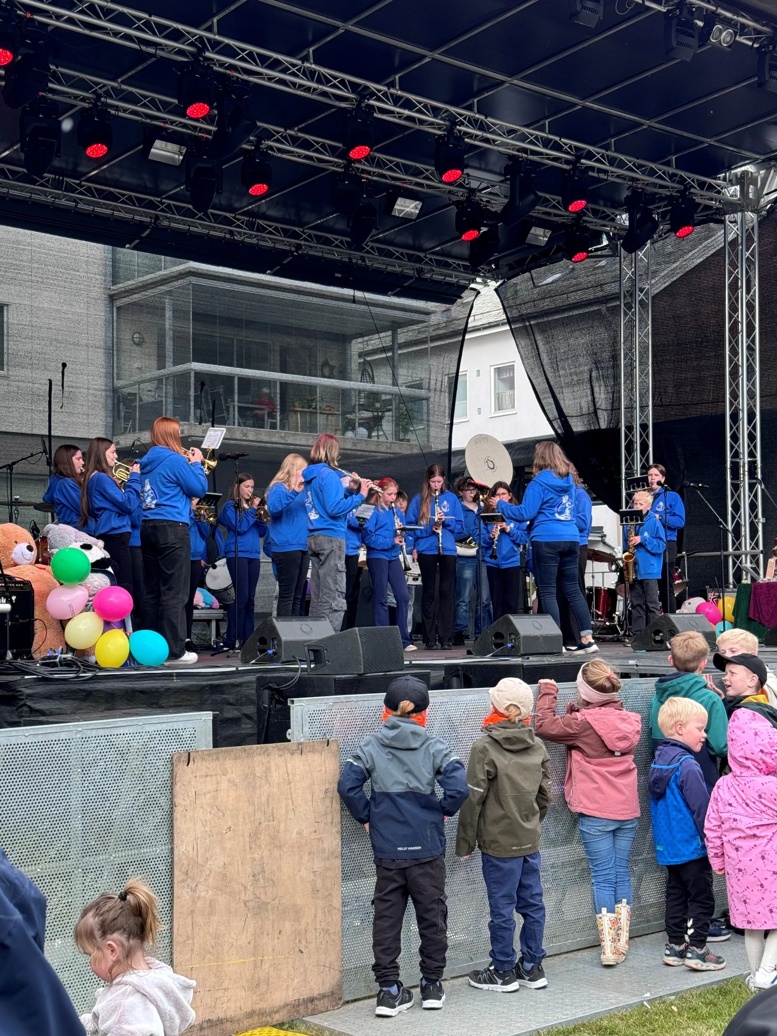 Barn og unge vender seg mot en utendørskonsert hvor en gruppe ungdommer spiller kobberinstrumenter på en scene. Det er ballonger på scenen, og publikum står foran, noen med barnevennlige klær og capser. Bygninger i bakgrunnen.